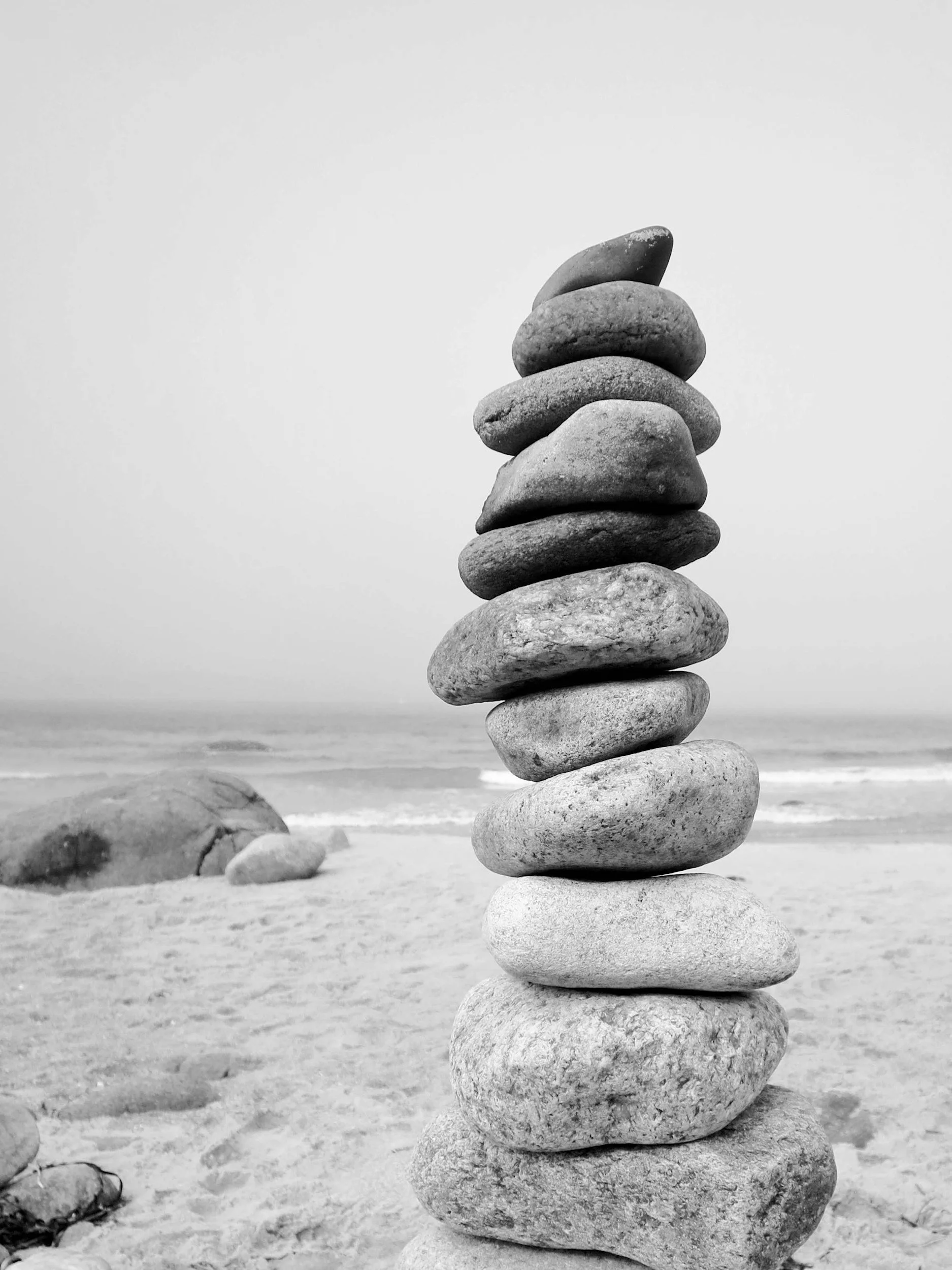 Stack of rounded stones on a sandy beach, with the ocean and cloudy sky in the background in black and white.