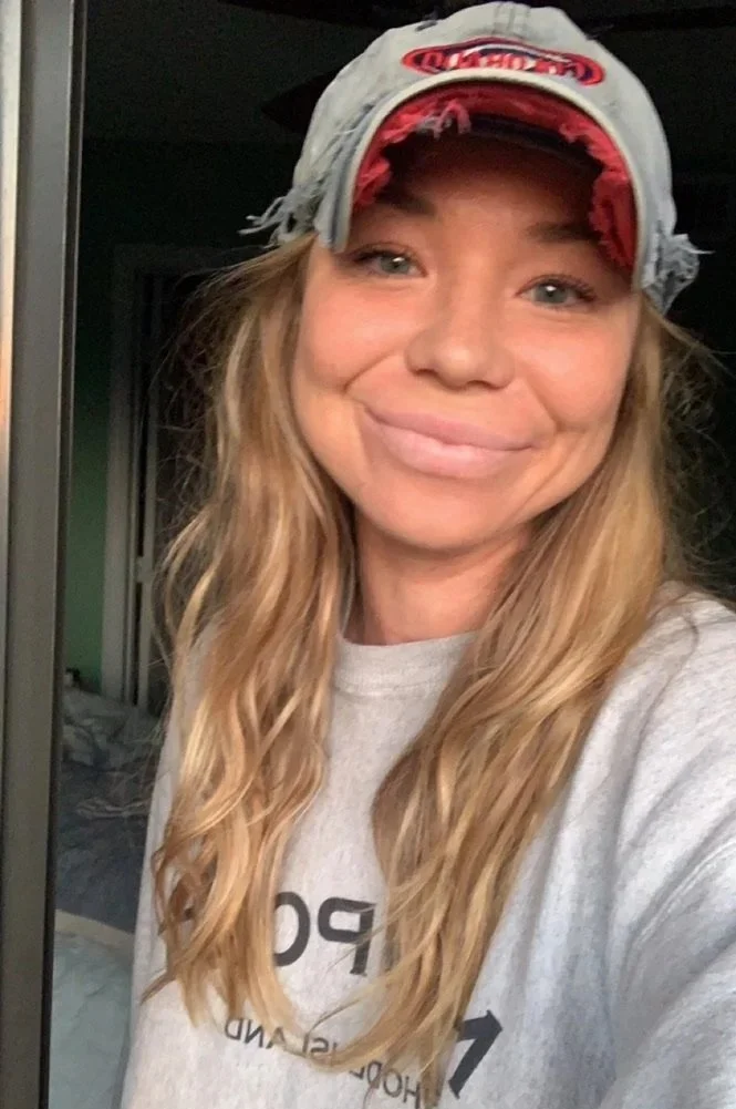 A woman with long, wavy blonde hair smiling in a casual indoor setting, wearing a gray baseball cap with red accents and a logo, and a light gray t-shirt.