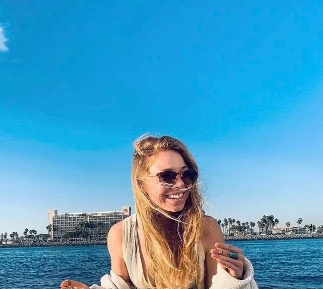 A smiling woman with sunglasses and long blonde hair, wearing a beige tank top, stands near the water with a cityscape and palm trees in the background on a sunny day.