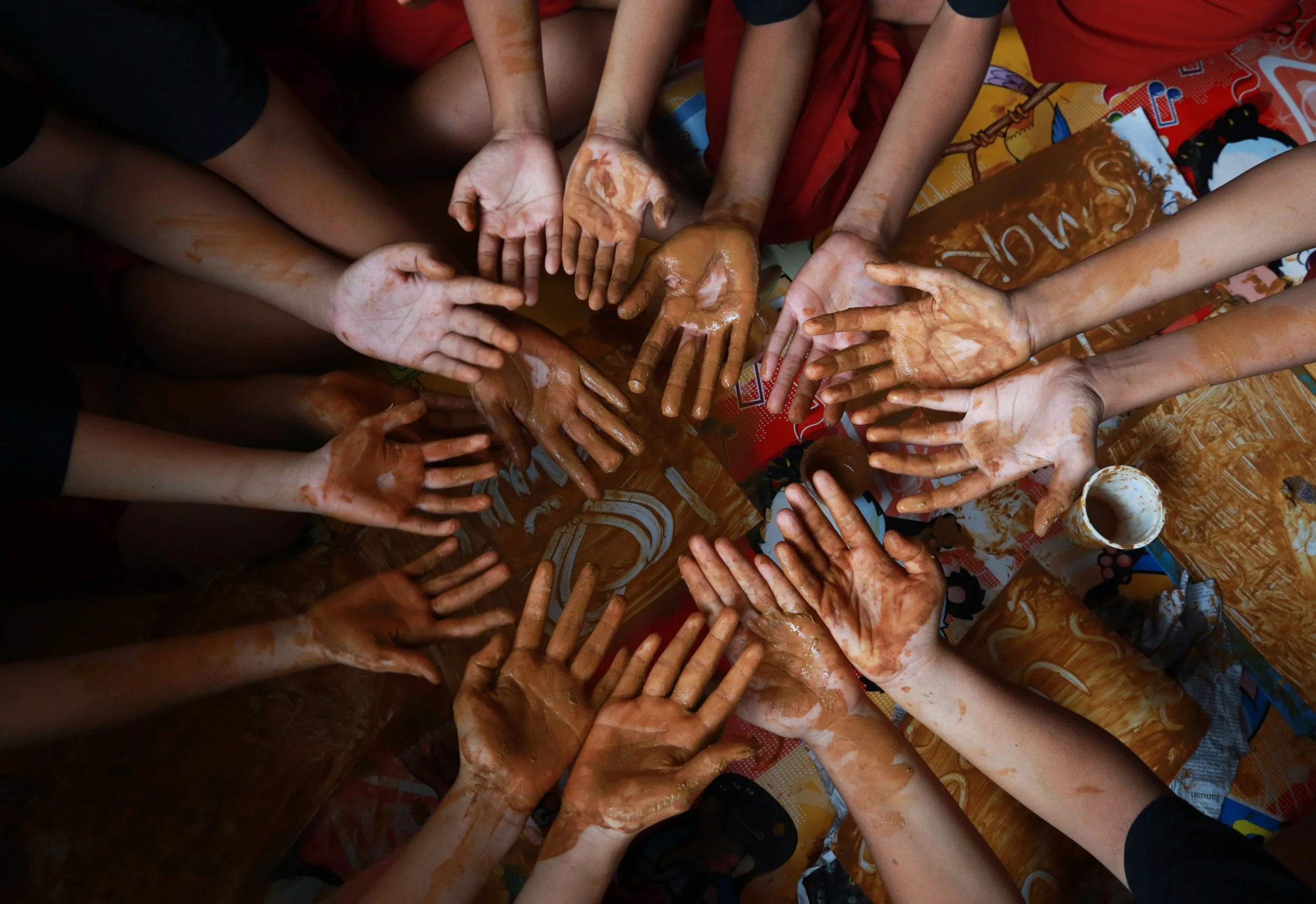 Multiple children with clay-covered hands reaching towards the center on a colorful table.