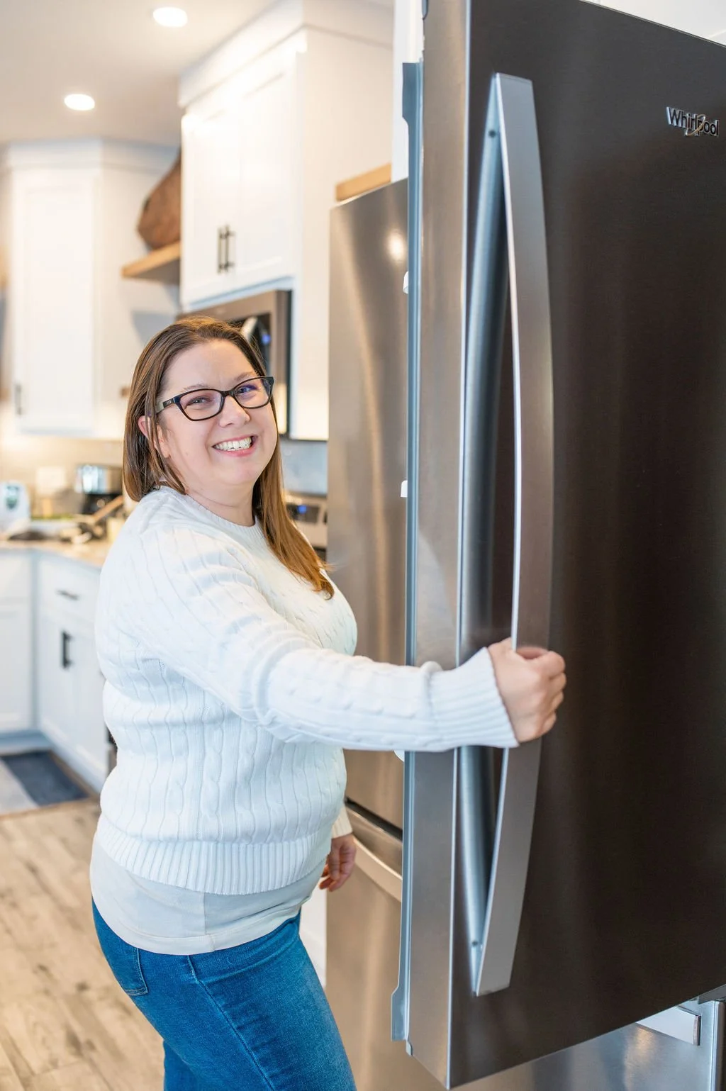 A smiling woman with glasses opening a stainless steel refrigerator door in a bright kitchen.
