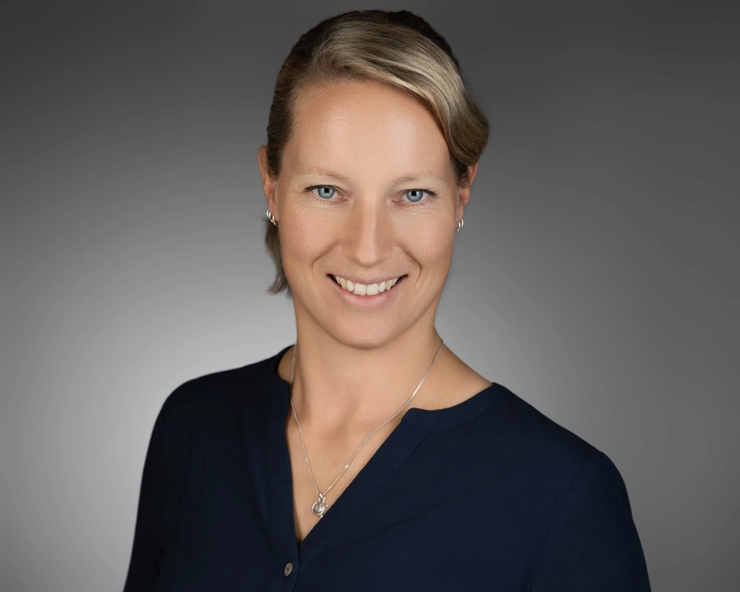 Smiling woman with short blonde hair and blue eyes, wearing a navy blue top and jewelry, against a gray background.