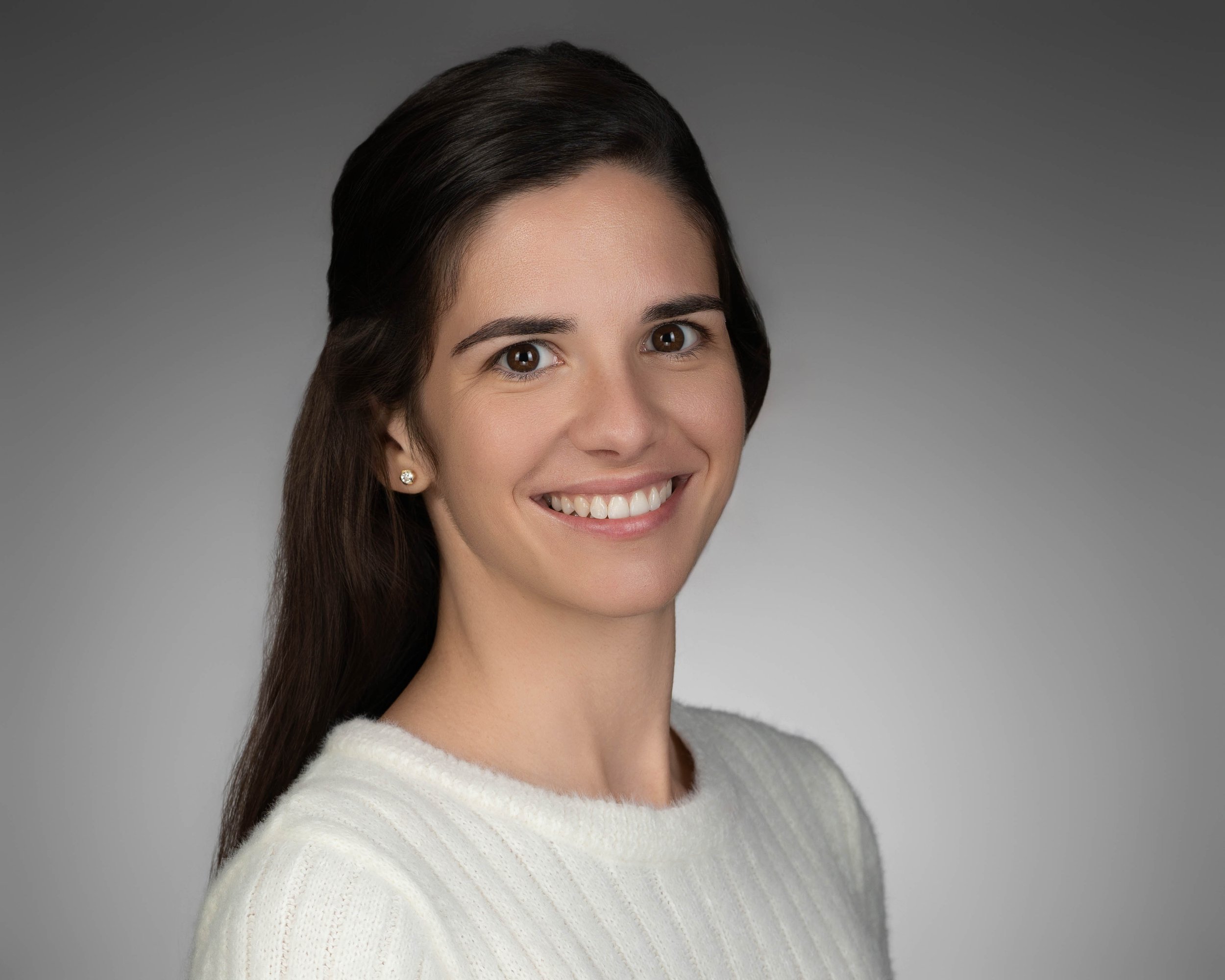 A young woman with long dark hair, wearing a white sweater and small earrings, smiling against a gray background.