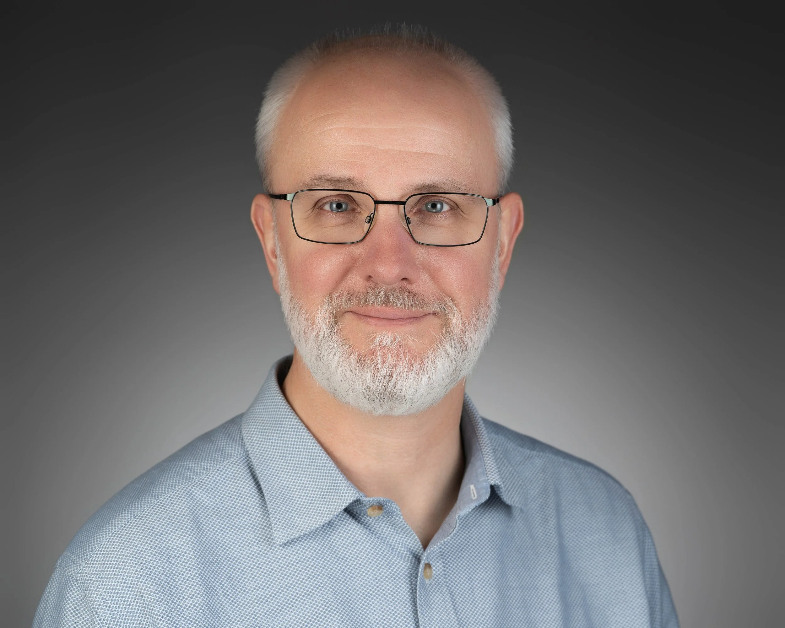 A middle-aged man with white hair, a beard, glasses, wearing a light blue collared shirt, dark background.