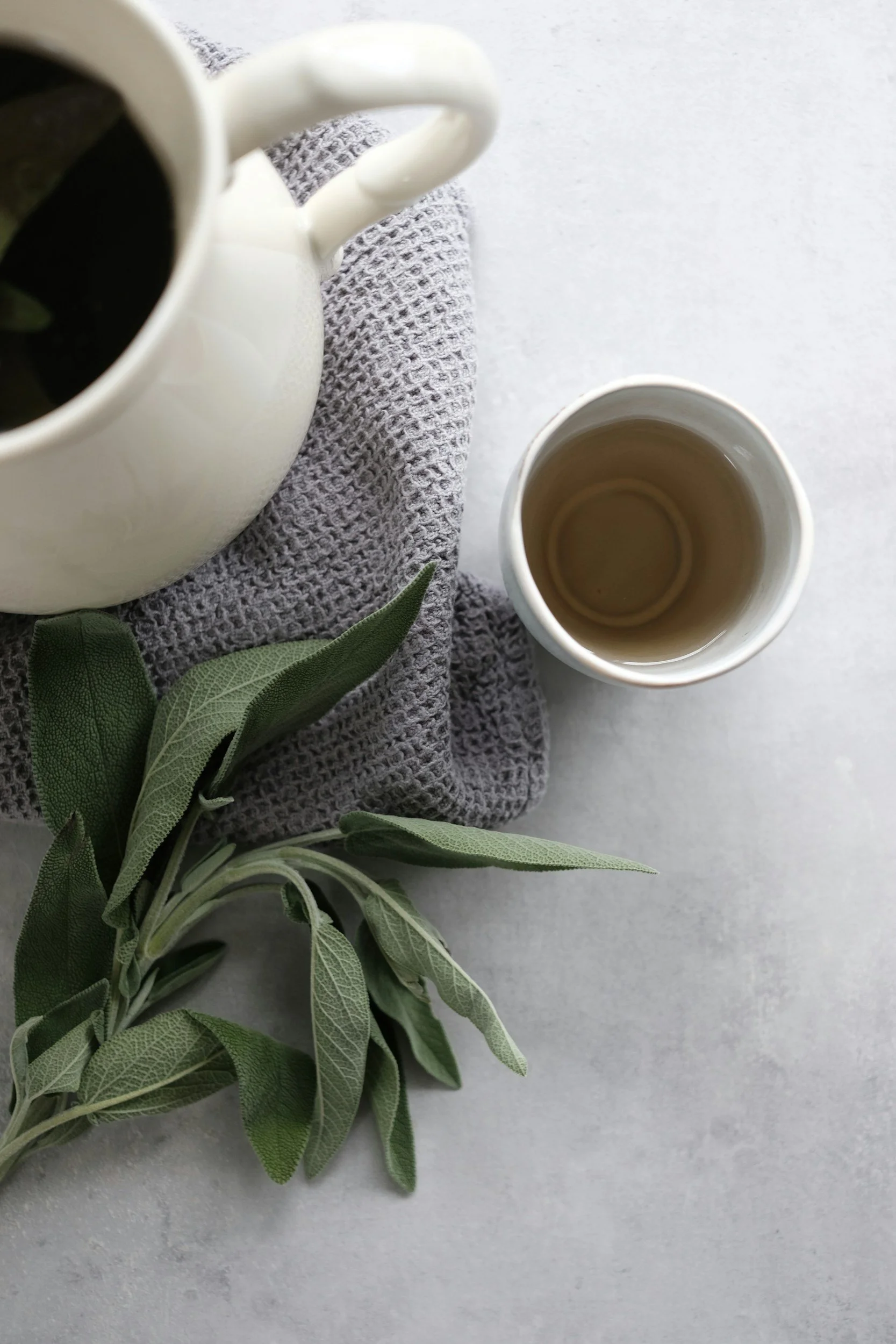 Top view of a white mug filled with black coffee next to a smaller white cup containing tea, placed on a light gray surface with a gray towel and green leaves.