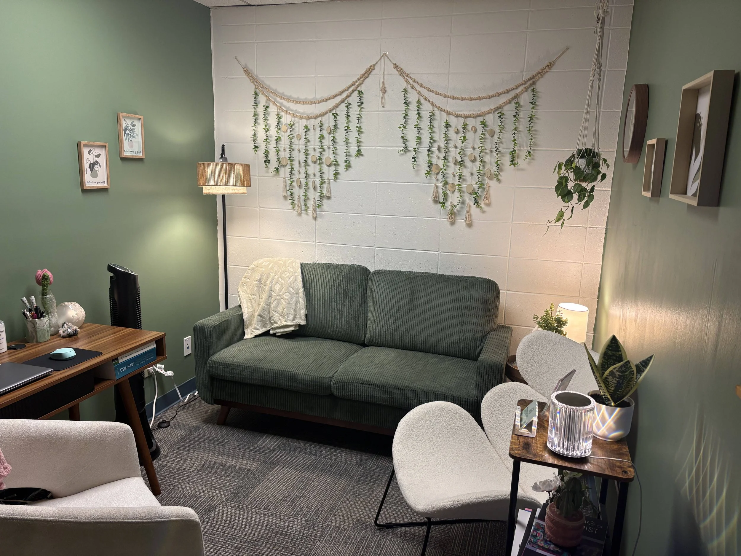 Cozy with green and white walls, featuring a green sofa with a beige throw, a wooden side table with a plant and candle, a white armchair, a desk with office supplies, a floor lamp, and decorative wall hangings including framed art and woven wall decor.