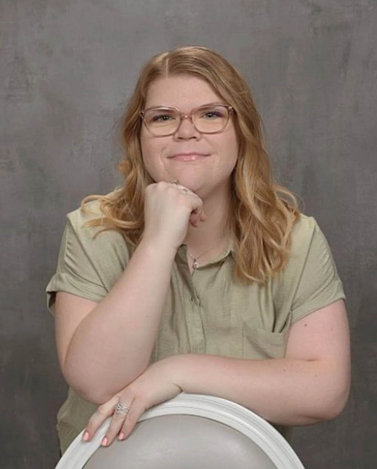 A woman with red hair, glasses, and a light green shirt resting her chin on her hand while leaning on a white chair.