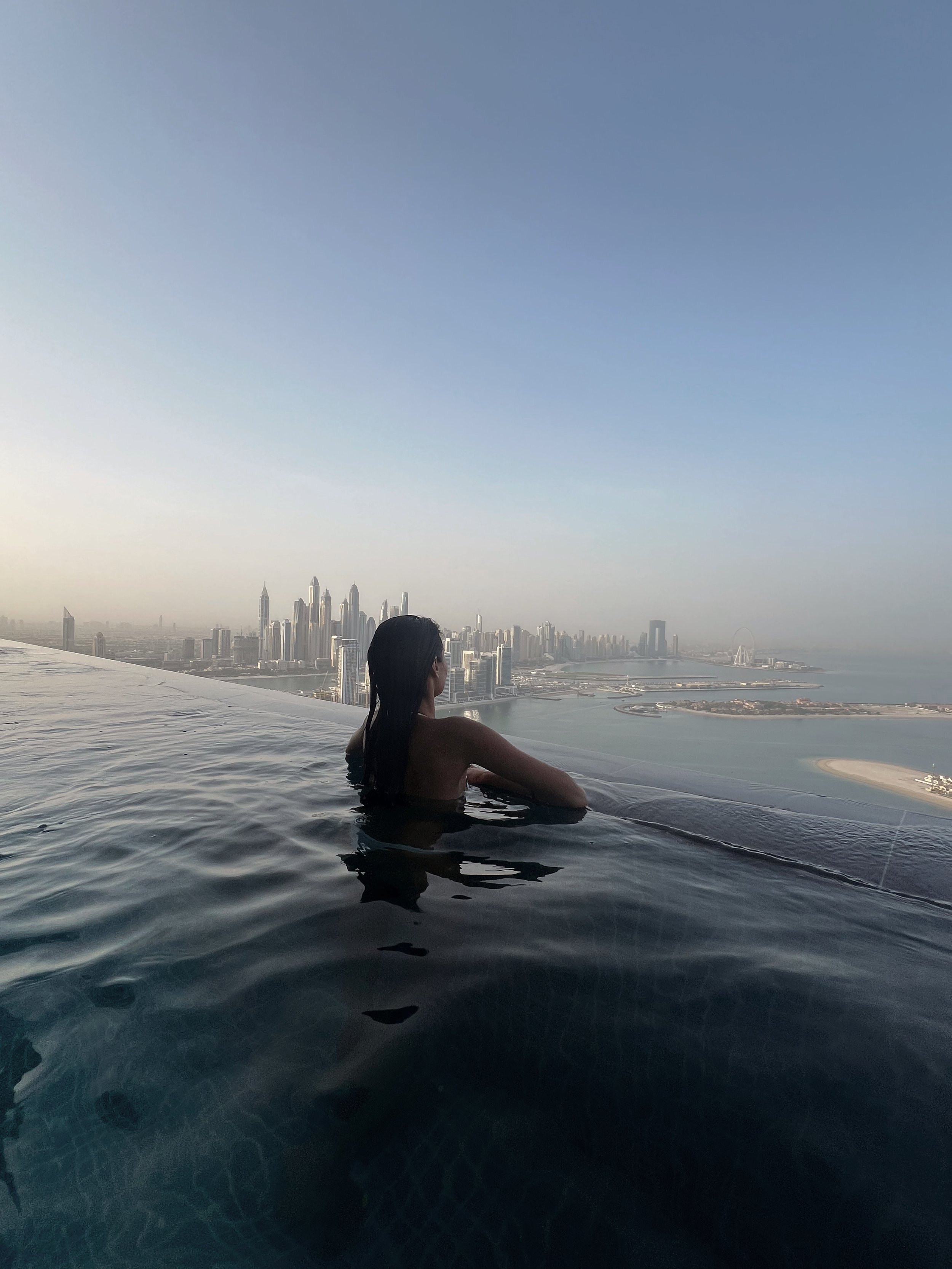 Woman with long dark hair relaxing in a rooftop infinity pool overlooking a city skyline including tall skyscrapers and a river, with clear blue skies.