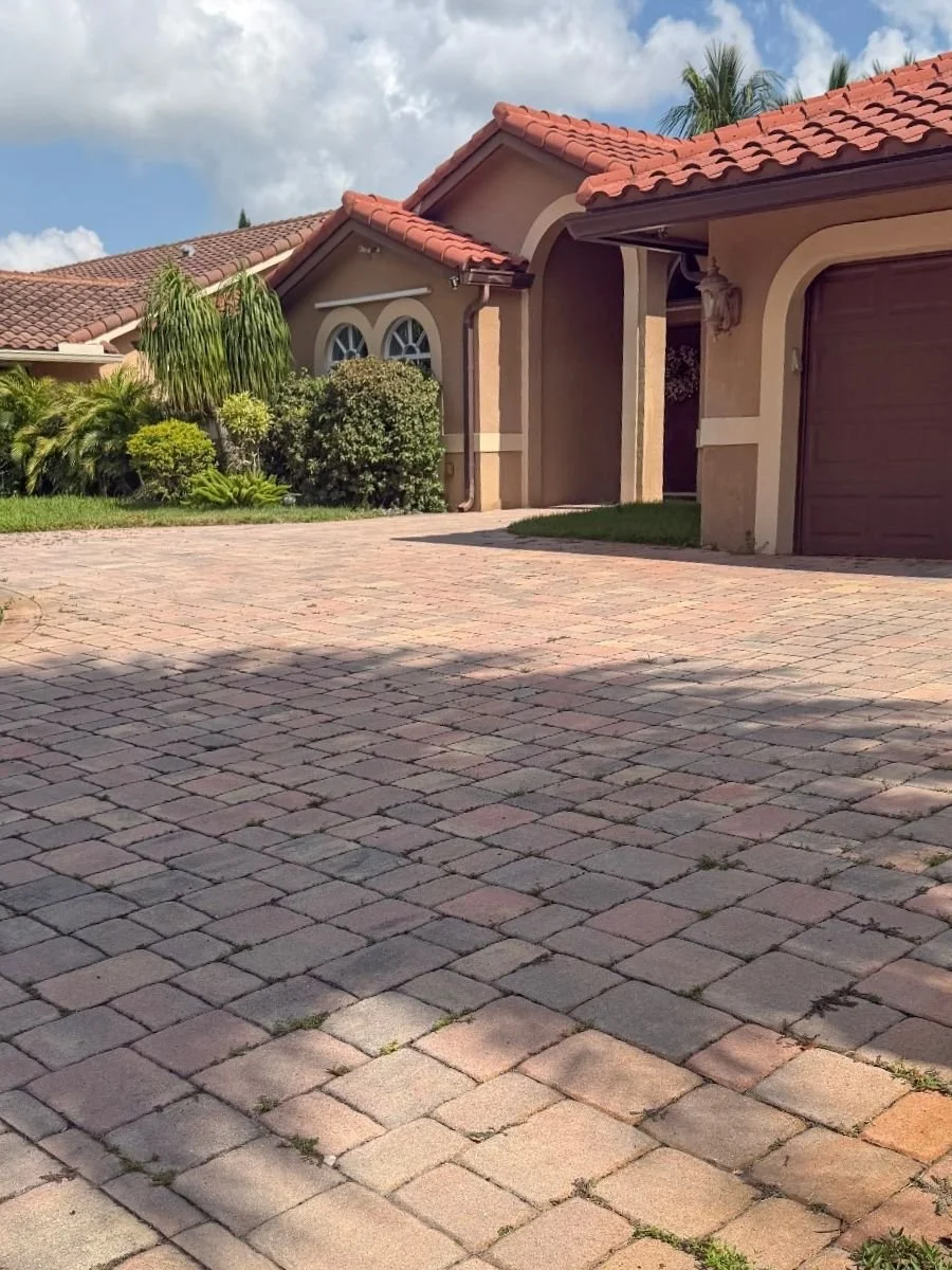 Residential brick paver driveway in Coral Springs before cleaning and sealing, showing weeds between joints and uneven surface color.