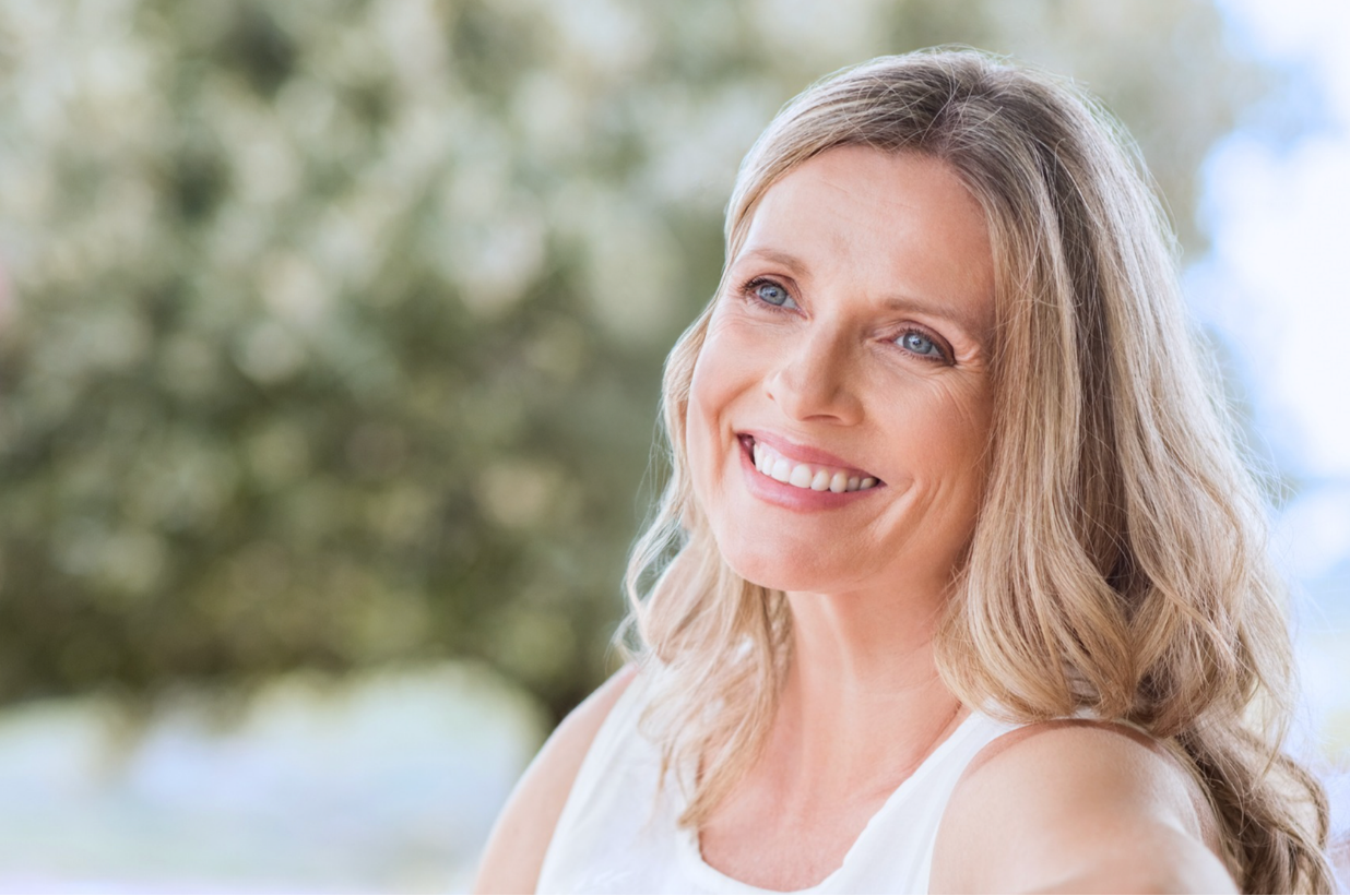 A smiling woman with blonde wavy hair, wearing a white sleeveless top, after personalized internal medicine in Austin TX at Westlake Medical Arts