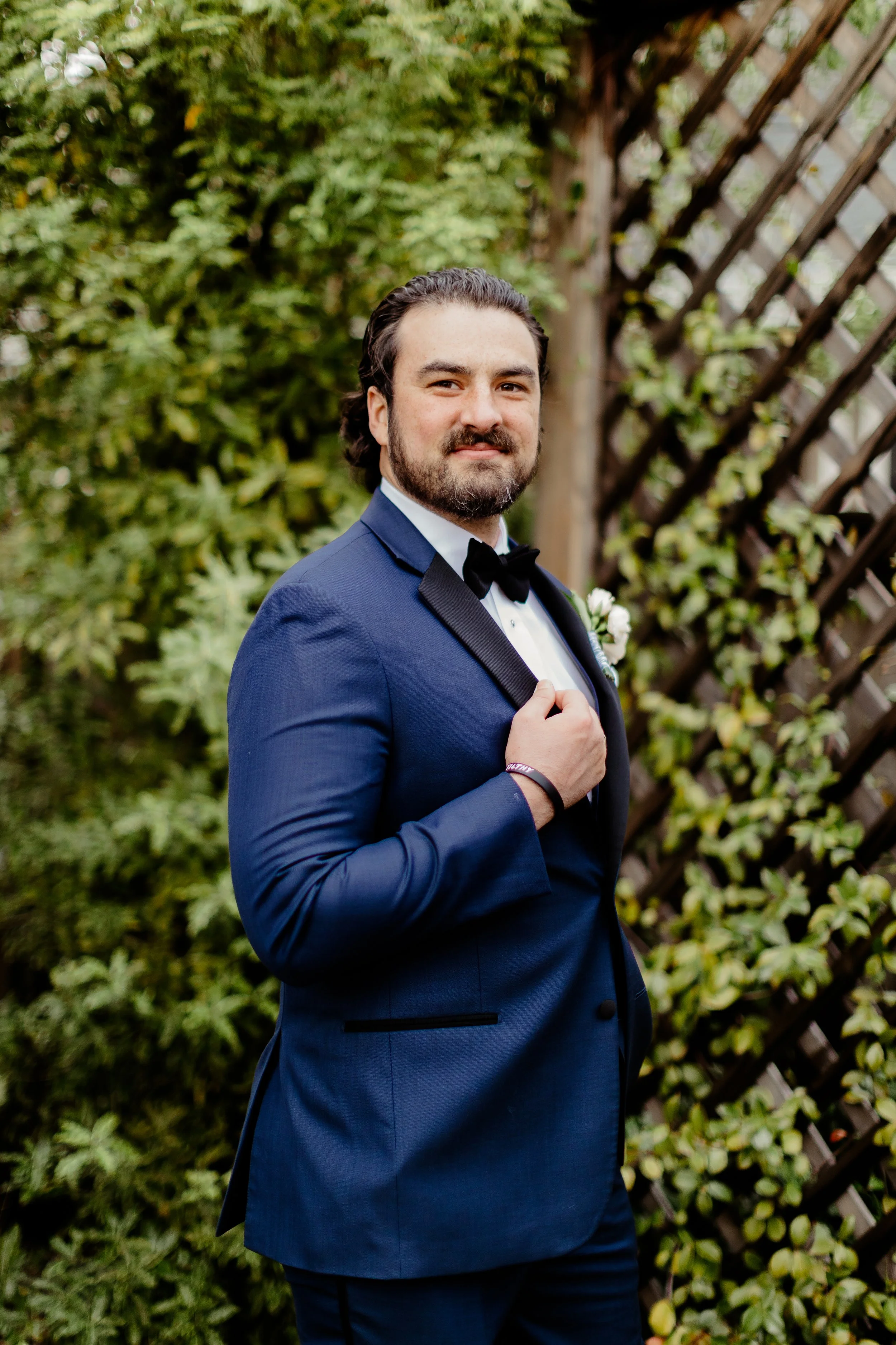 A man with dark hair and a beard wearing a dark blue tuxedo with a black bow tie, standing outdoors next to a wooden lattice and green foliage, holding the lapel of his tuxedo with his right hand.