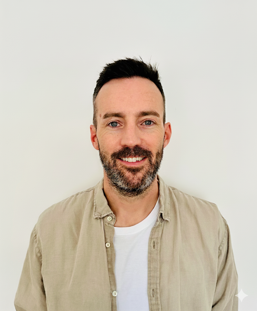 A smiling man with short dark hair, a beard, wearing a beige button-up shirt over a white t-shirt, standing against a plain white wall.