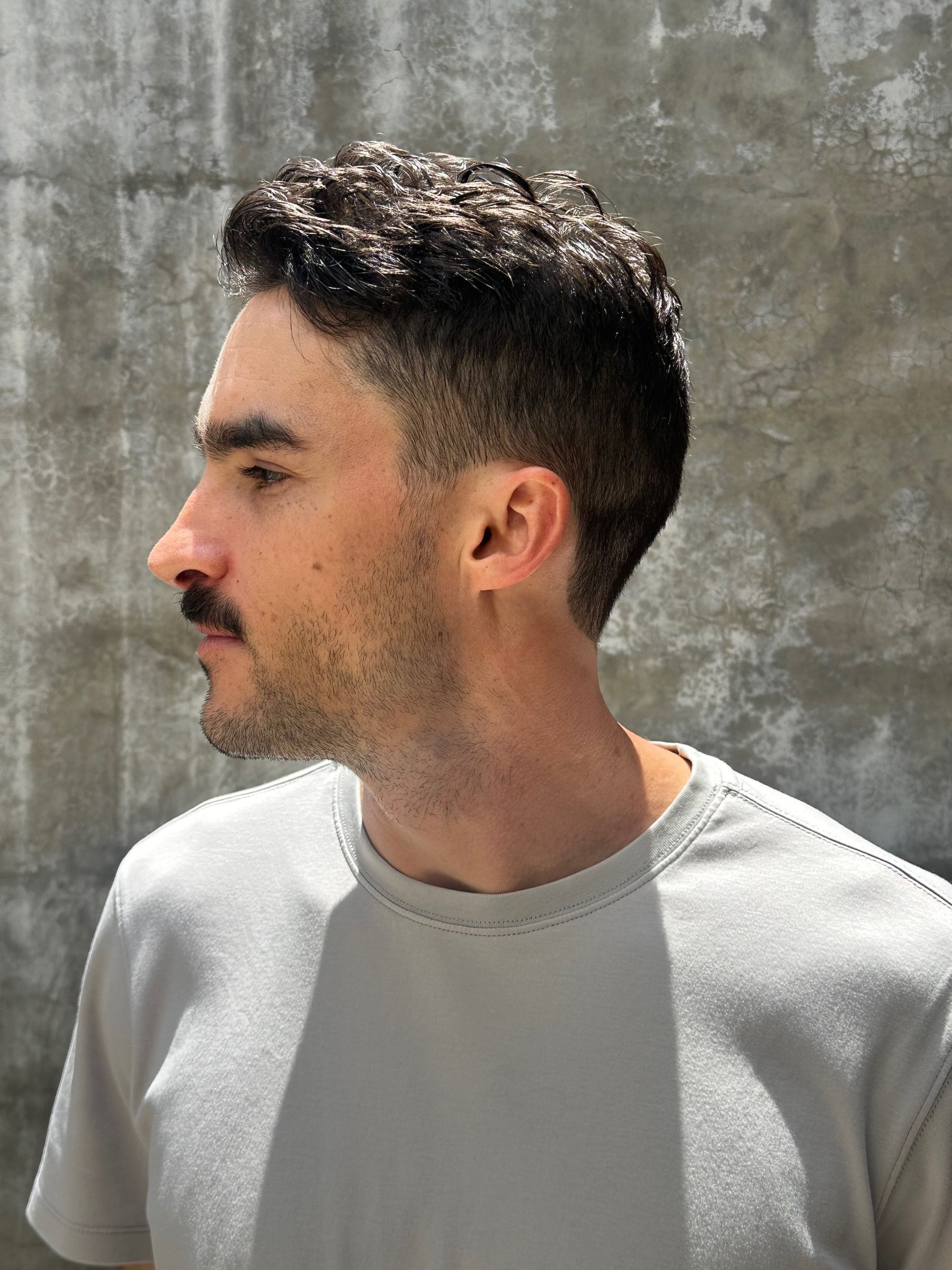 Side view of a man with short, dark, wavy hair and a light beard, wearing a light gray t-shirt, standing against a textured gray wall.