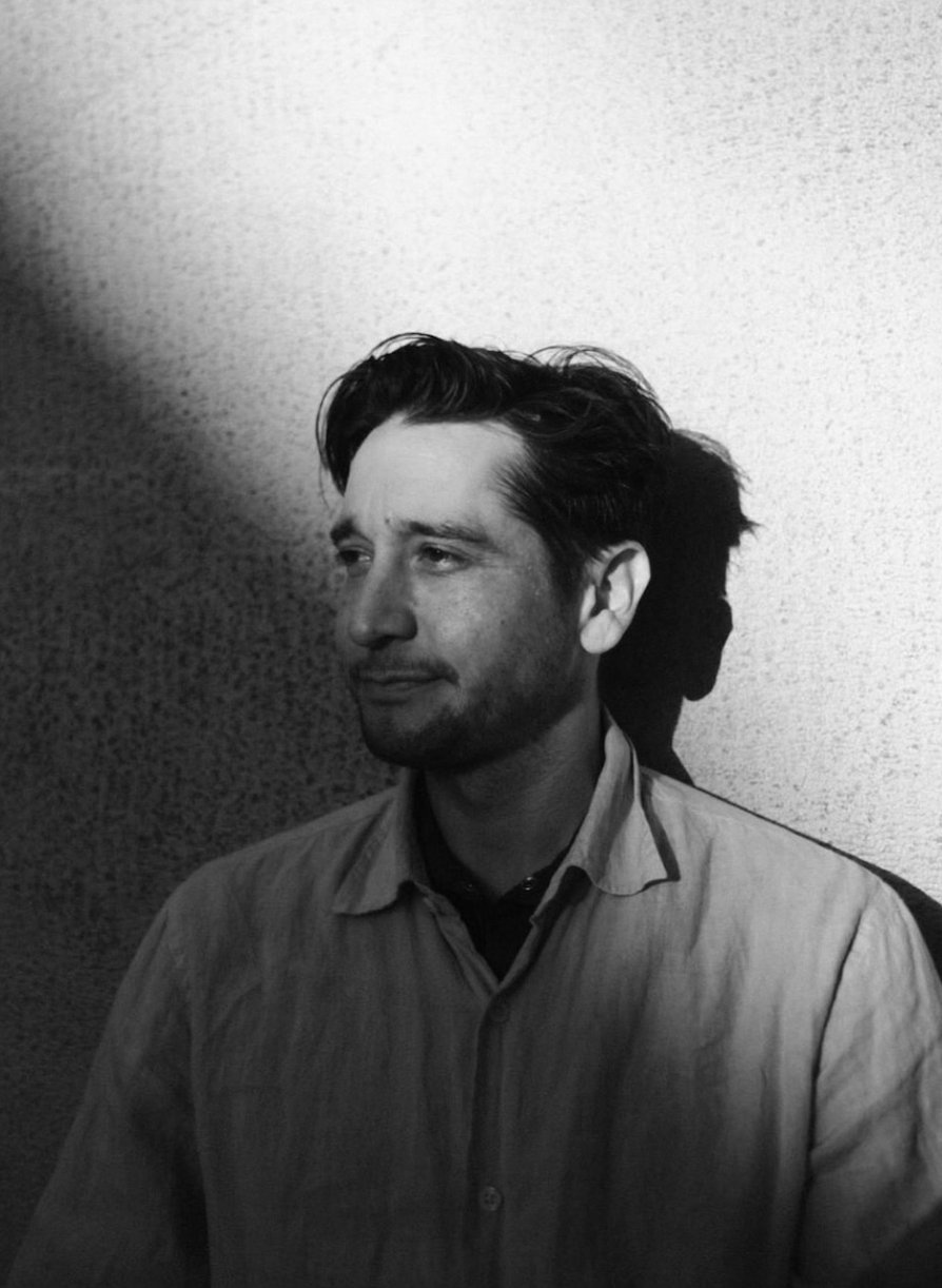 A black and white portrait of a young man with dark hair, wearing a collared shirt, standing against a textured wall, with shadow on the left side of his face.