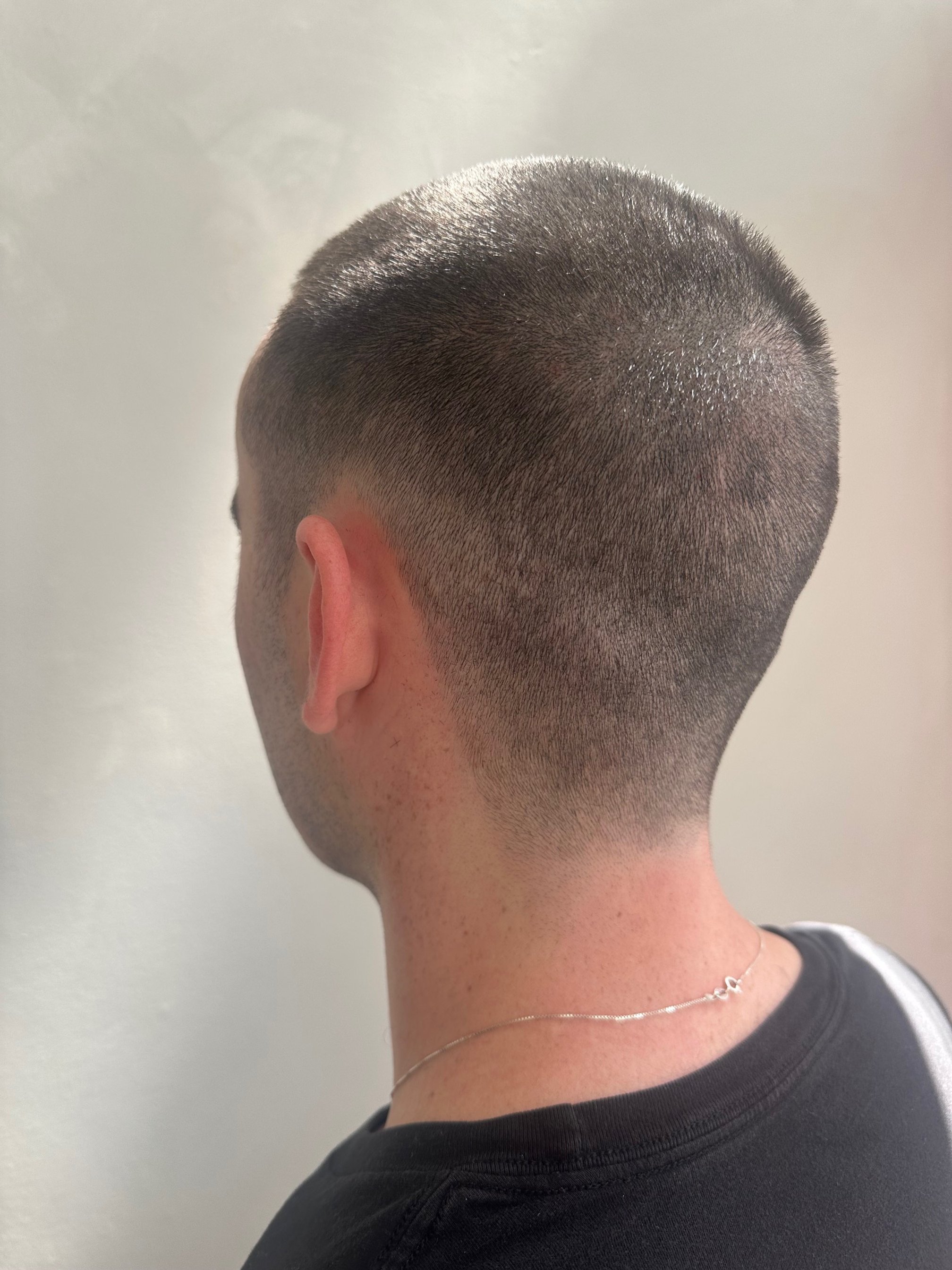 Back of a person's head with a close-cropped haircut, dark hair, and a neck tattoo, wearing a black shirt and silver chain necklace.