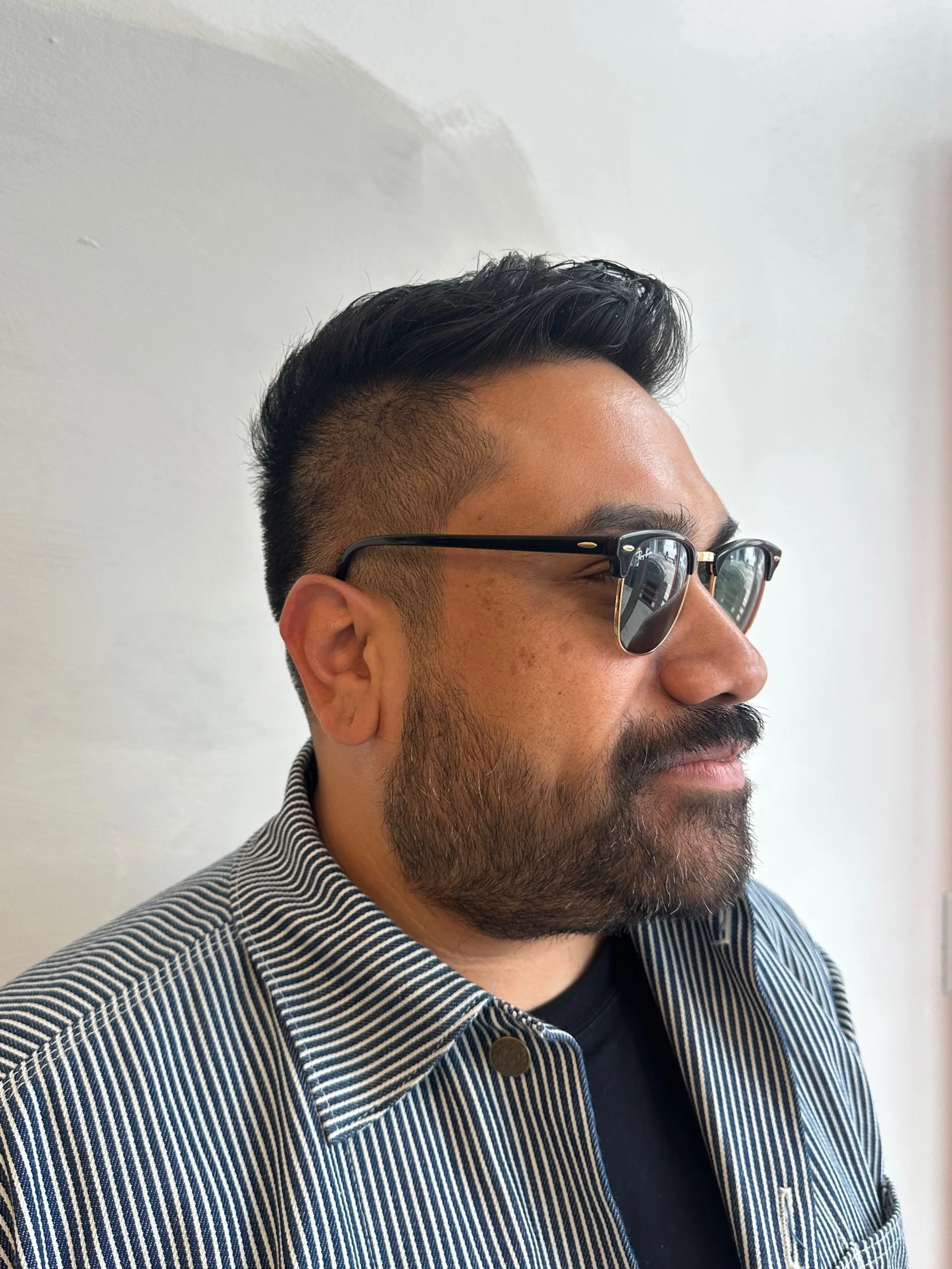 Side profile of a man with styled black hair, beard, wearing sunglasses with a reflection, and a striped shirt against a light background.