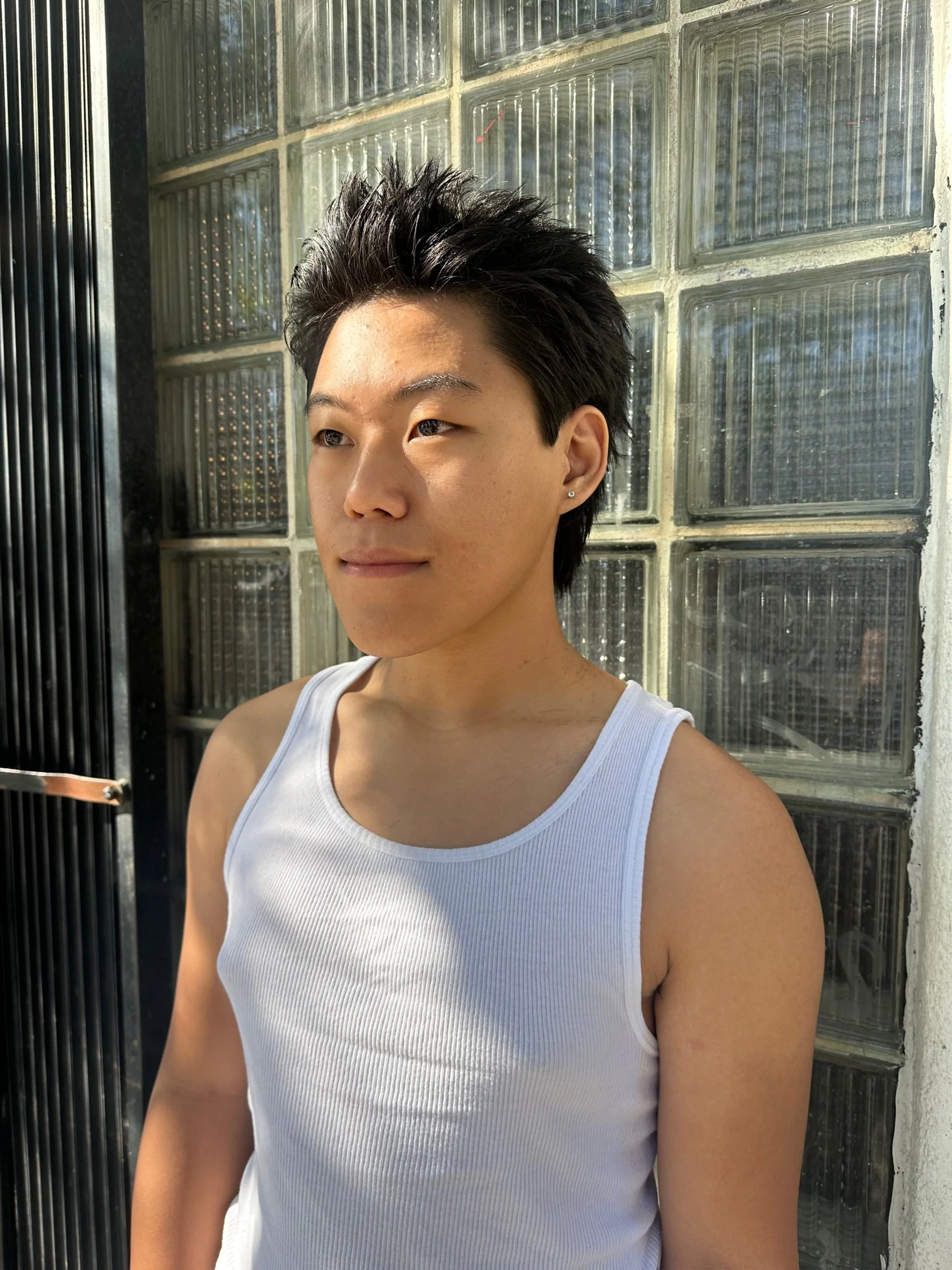 A young man with short, dark, spiky hair, wearing a white sleeveless shirt, is standing outdoors next to a glass block wall. The sun is shining on his face, creating shadows and highlights.