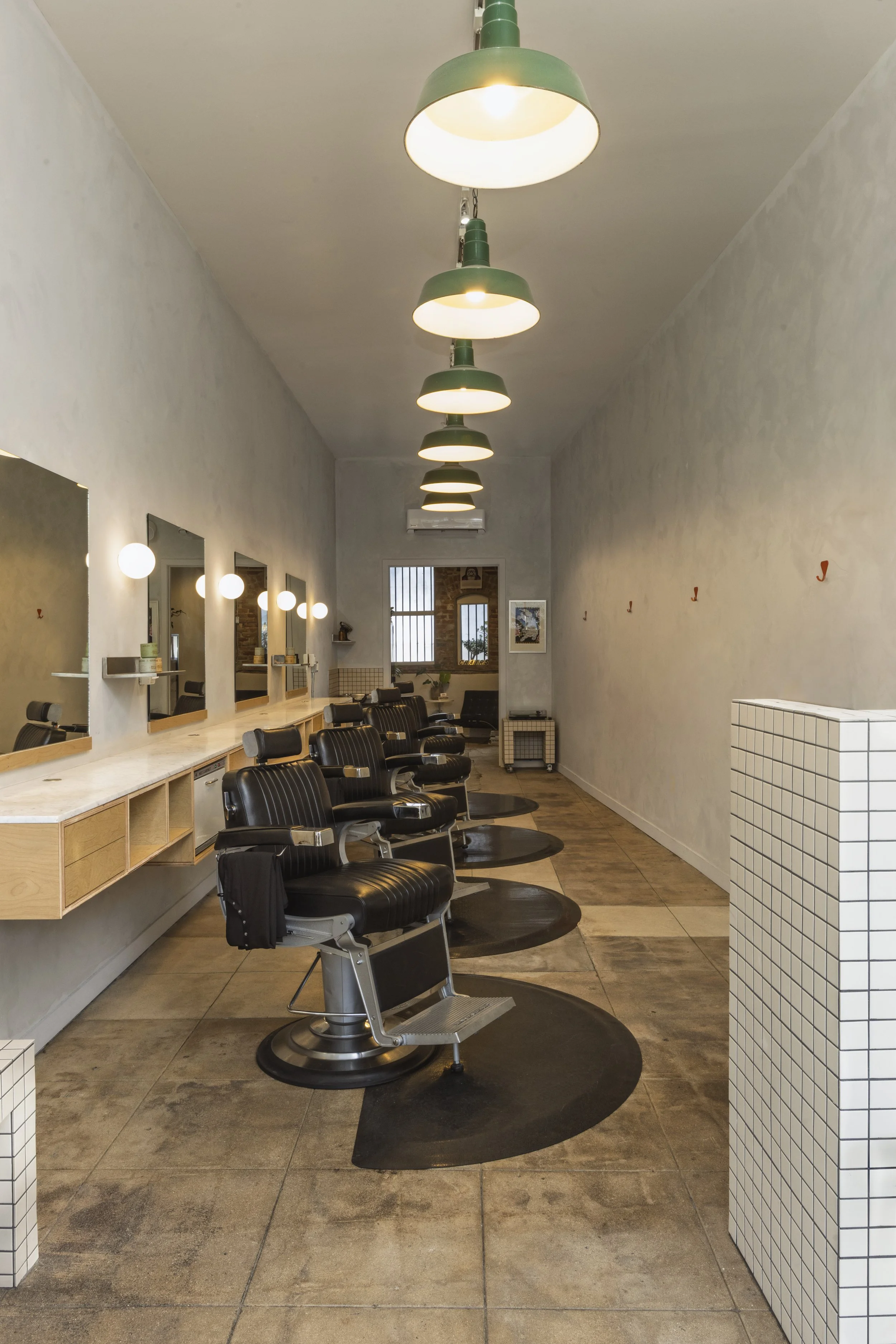Interior of a modern barbershop with four vintage-style black barber chairs, large mirrors, wooden shelves, green ceiling lights, and a tiled white partition wall.