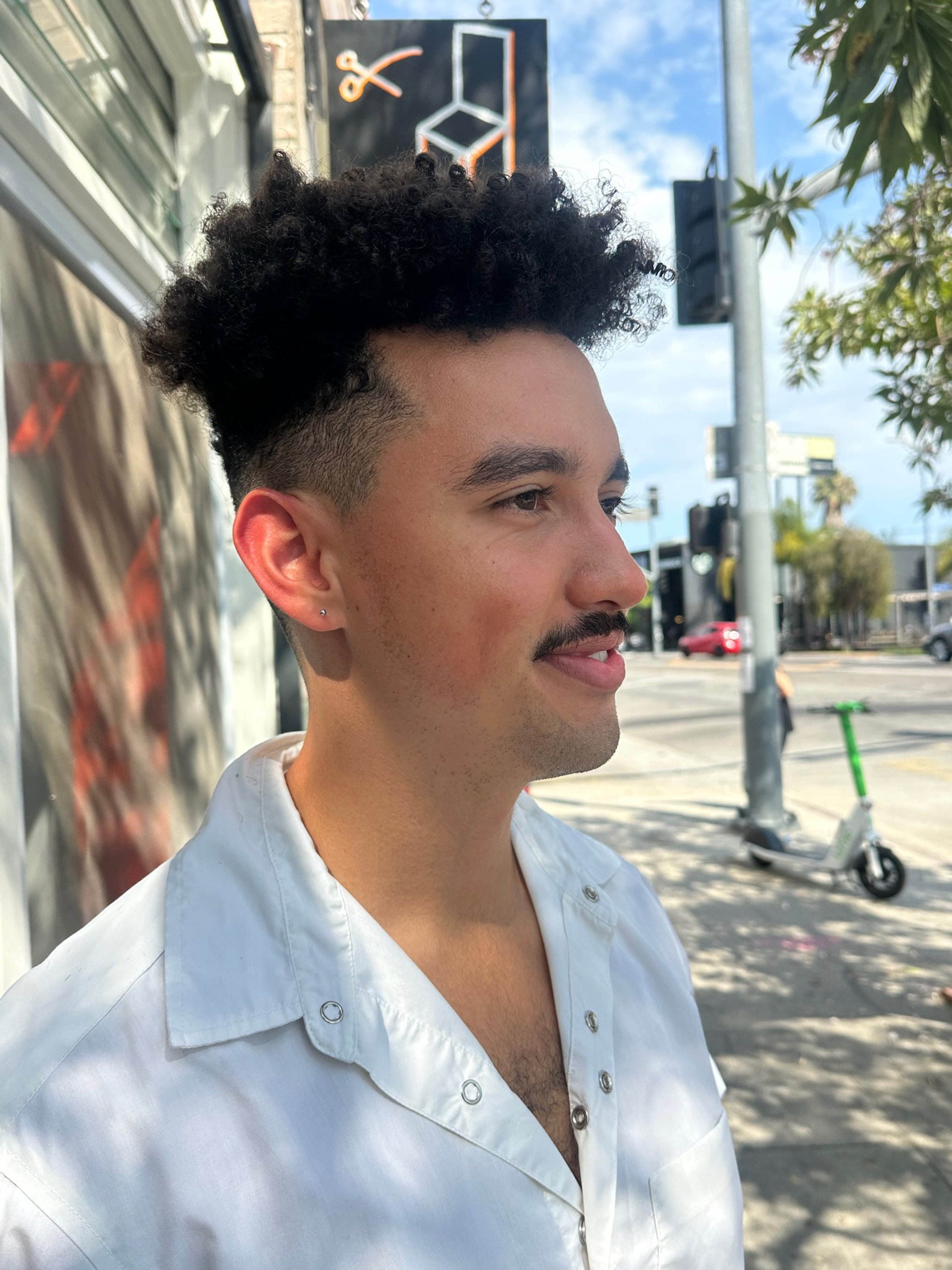 A young man with curly black hair and a mustache, wearing a white shirt, stands outdoors on a sunny day, with a street and traffic light in the background.