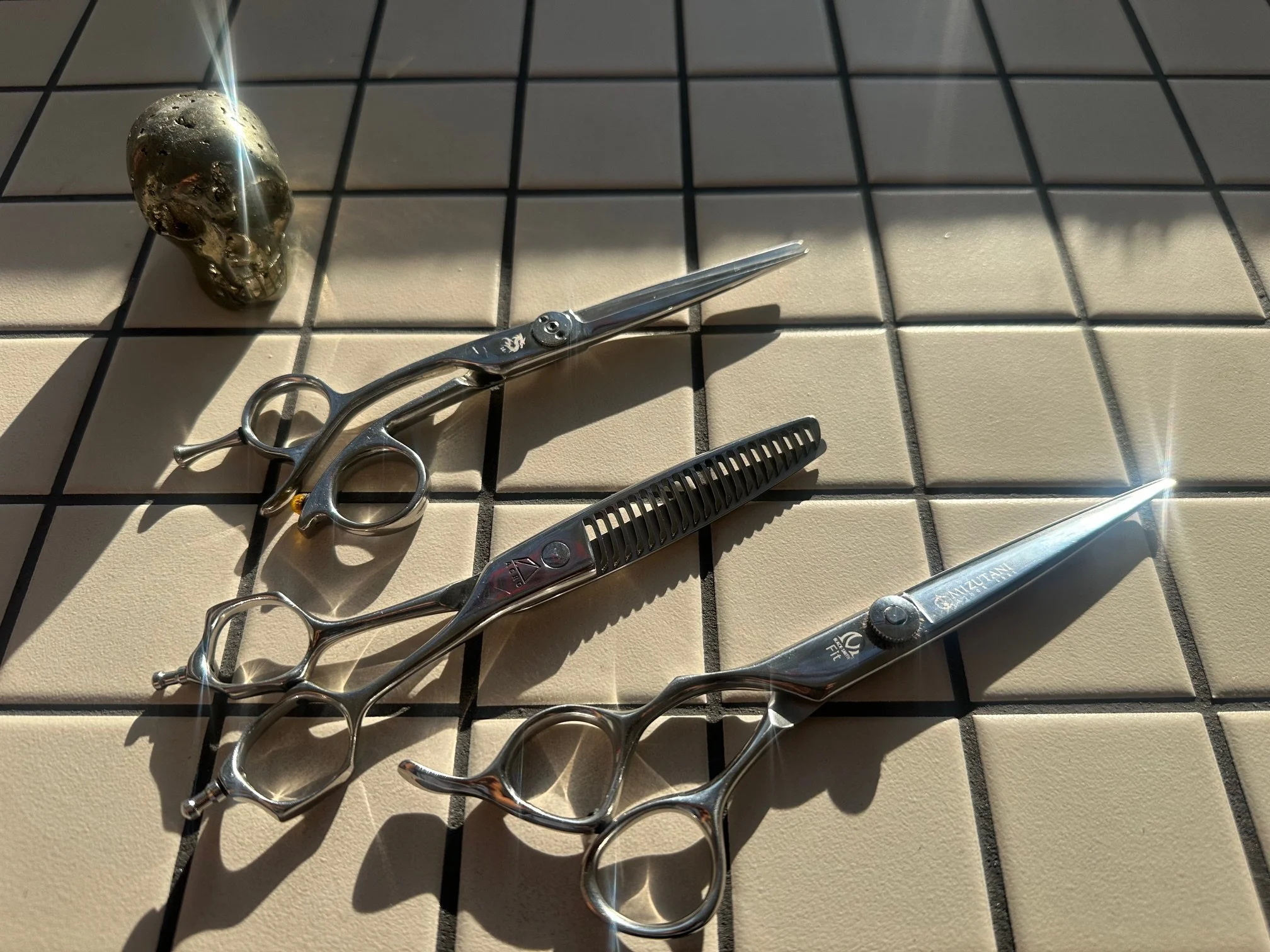 Three pairs of hair cutting scissors on a tiled surface with sunlight reflection and a decorative skull at the top left.