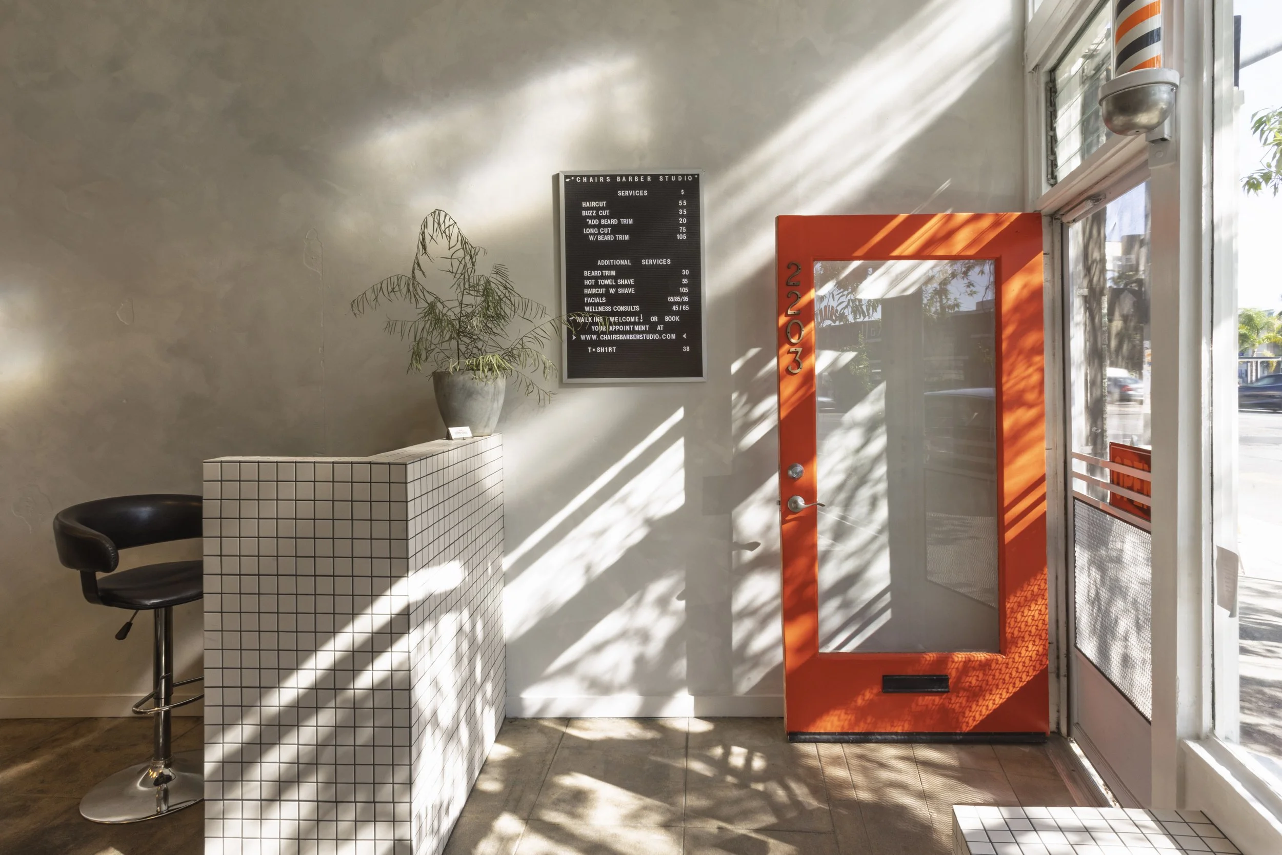 Interior of a barbershop with a white tiled reception counter, a black bar stool, a potted plant on the counter, a black menu board on the wall listing services and prices, an orange barber shop pole sign, and a glass door with sunlight and shadows cast on the wall.
