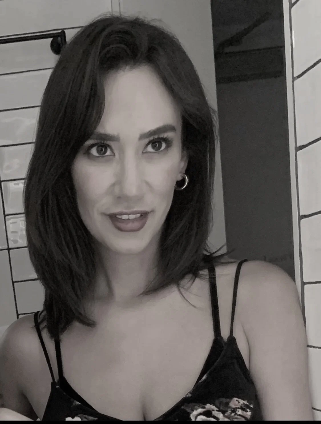 A woman with shoulder-length brown hair, wearing makeup including red lipstick and hoop earrings, smiling at the camera in front of tiled walls.