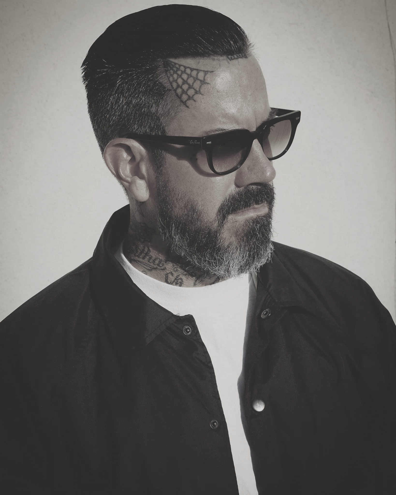 A man with a beard and tattoos wearing sunglasses and a dark jacket, standing against a light-colored background.