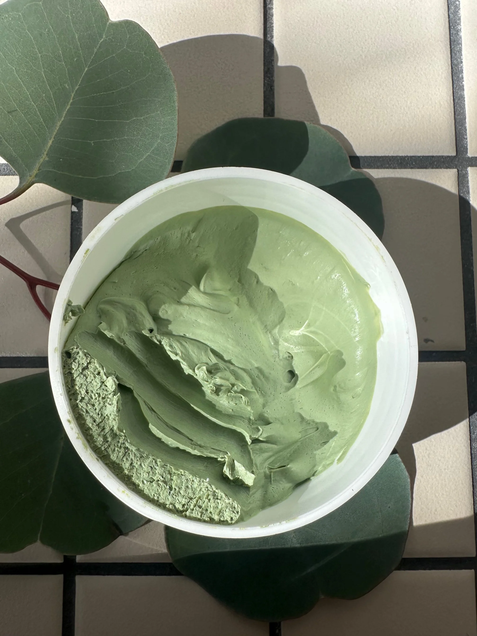 A bowl of green matcha and tulsi face mask on a tiled surface surrounded by green eucalyptus leaves.