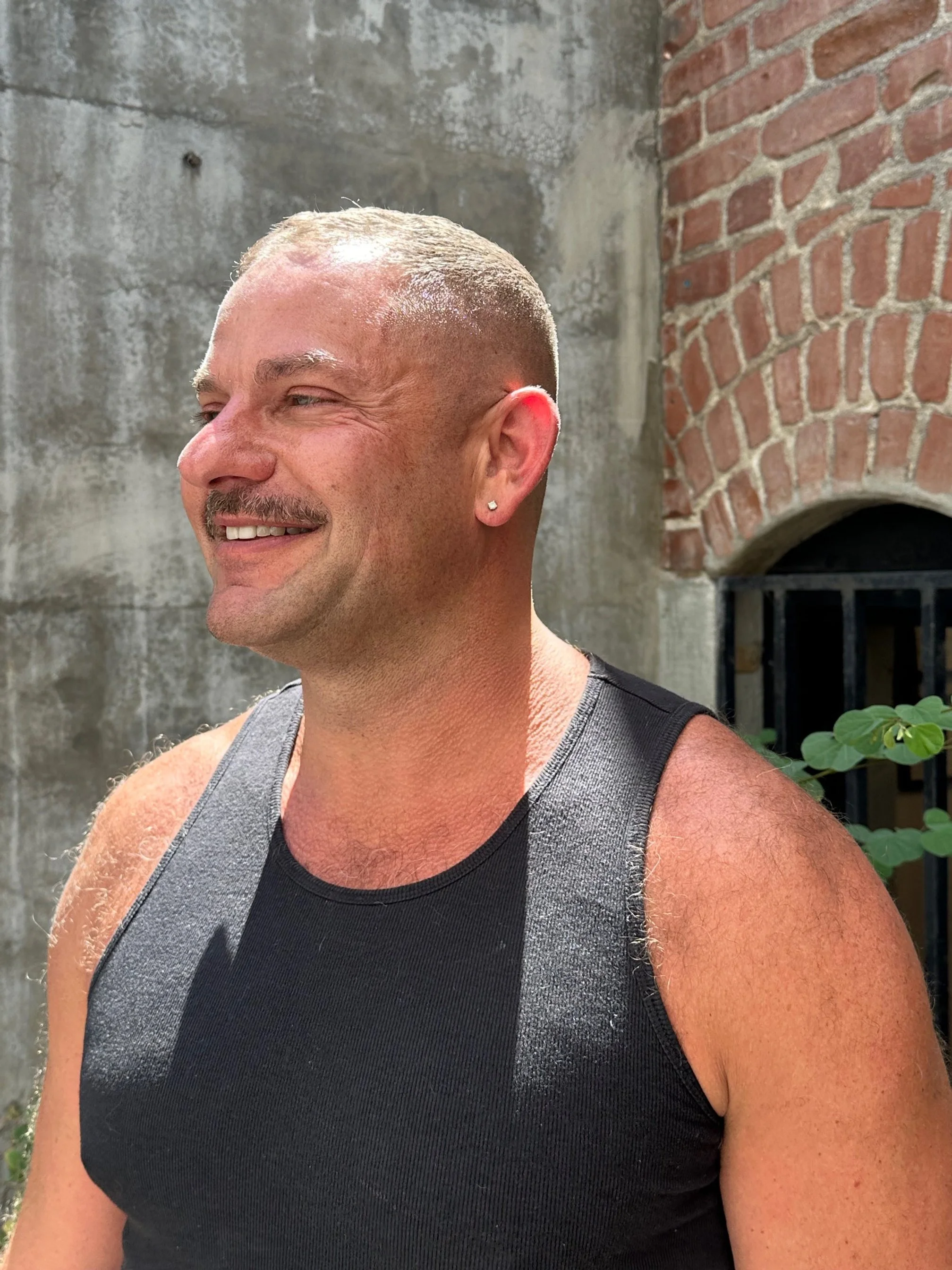 A smiling man with a buzz cut hairstyle, wearing a black sleeveless athletic shirt and a small earring in his left ear, standing outdoors against a background of a textured concrete wall, a brick structure, and some greenery.