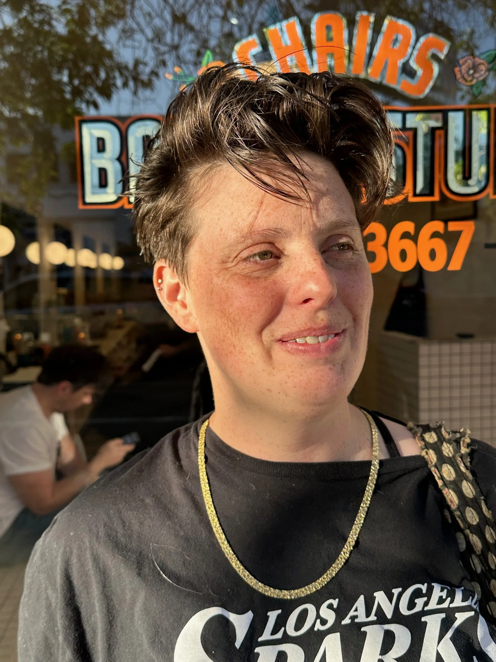 A smiling person with short, spiky hair and a black T-shirt with white lettering, standing outside in front of a window with a colorful sign, during early evening or sunset.