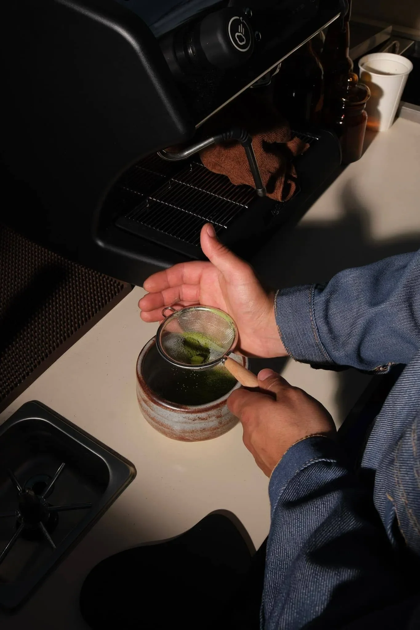 Person pouring a green liquid from a small tea strainer into a ceramic cup on a white countertop, with a coffee machine and various containers in the background.