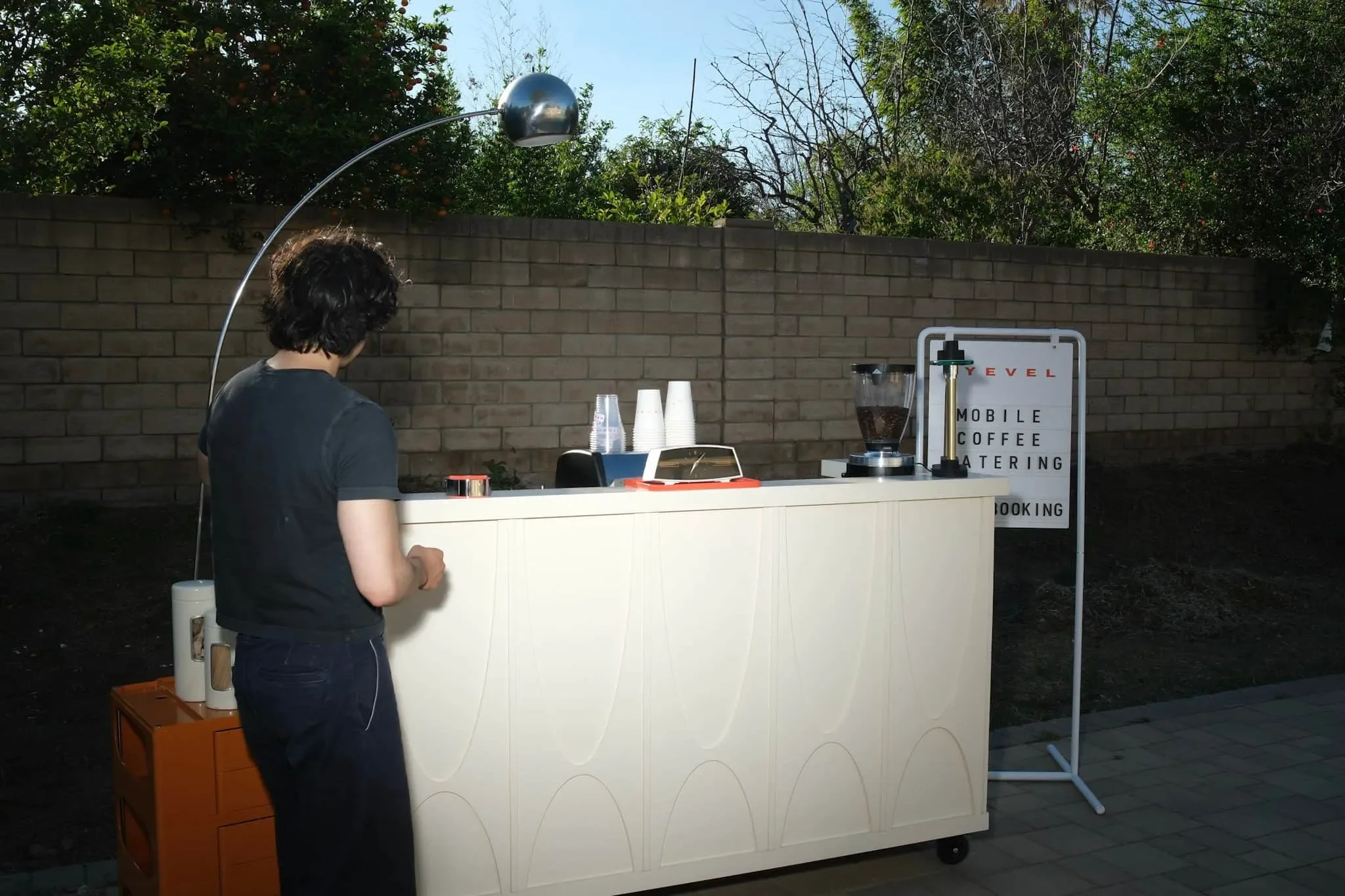 A person with dark curly hair wearing a black shirt stands in front of a white mobile coffee catering stand outdoors. The stand has three stacked disposable cups, a coffee grinder, and a sign that reads 'Mobile Coffee Catering.' There is a small oran
