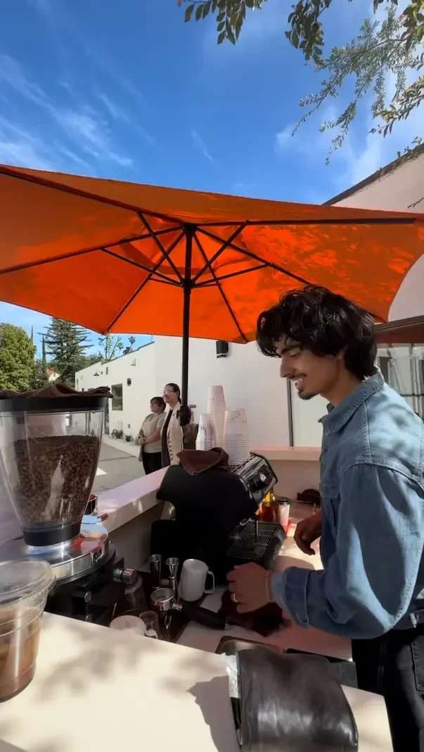 A barista making coffee at an outdoor cafe counter under an orange umbrella, with a clear blue sky overhead and a few people standing in the background.