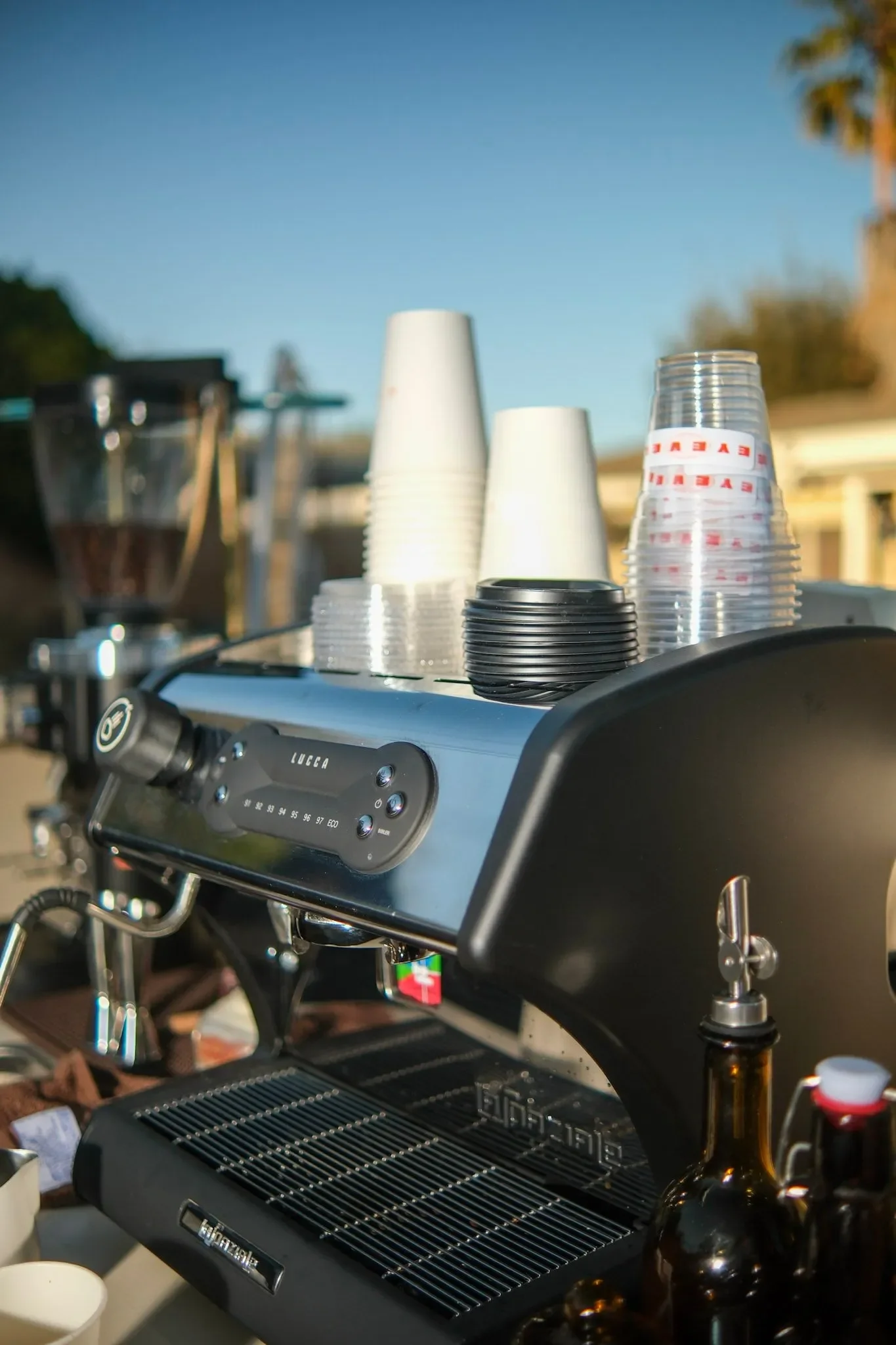 Close-up of a professional espresso machine with stacked disposable cups, a glass jar, and a bottle placed on it during daytime, outdoors, with trees and buildings in the background.