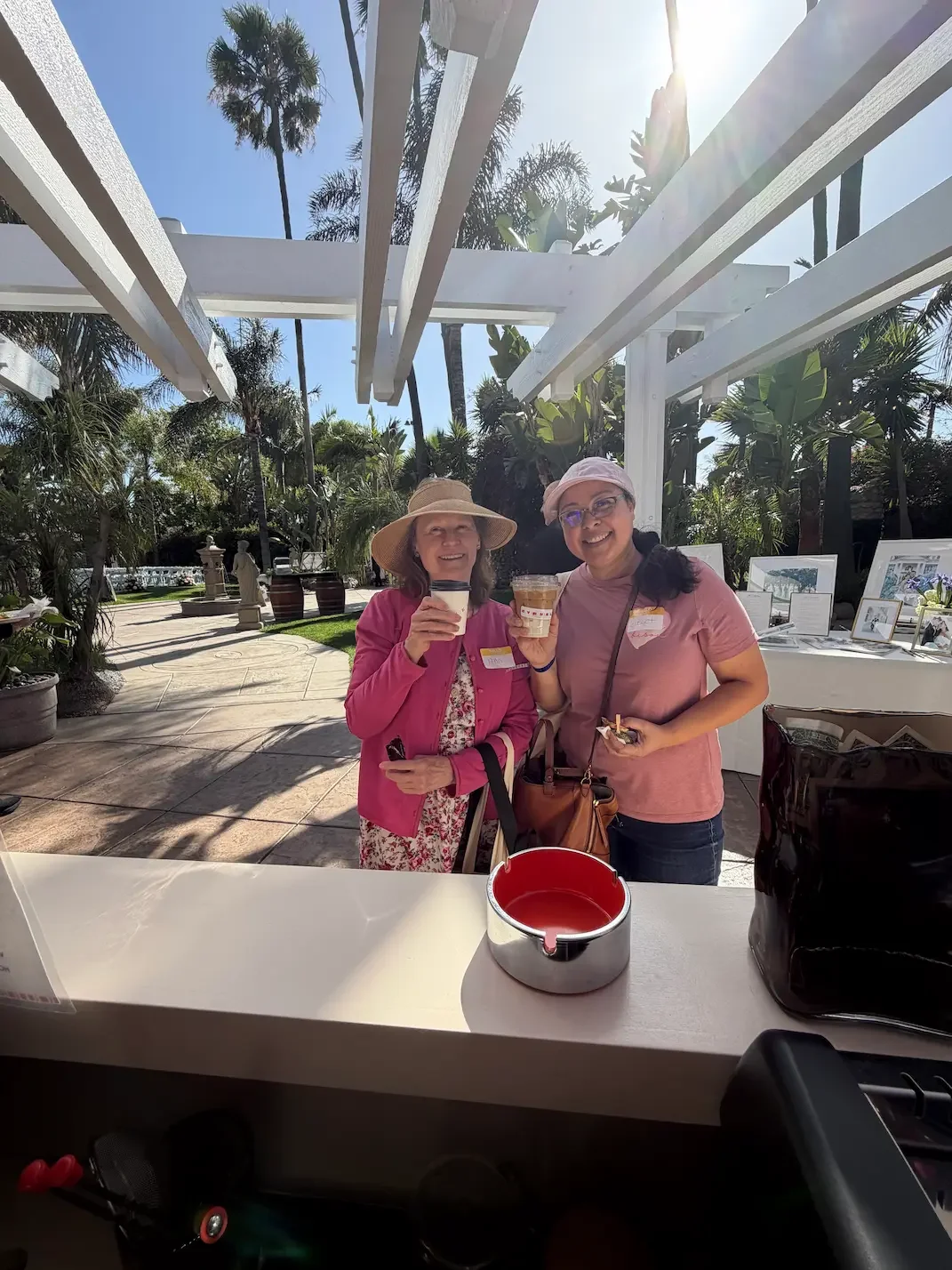 Two women smiling and holding drinks in an outdoor setting with palm trees and bright sunlight.