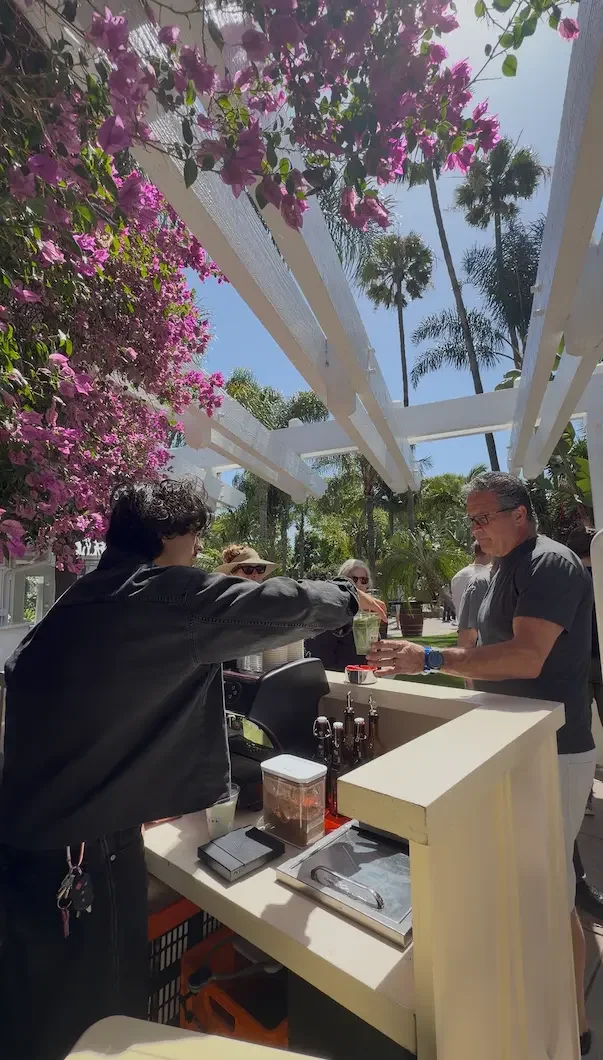 People at an outdoor café counter with pink bougainvillea flowers and tall palm trees in the background on a sunny day.
