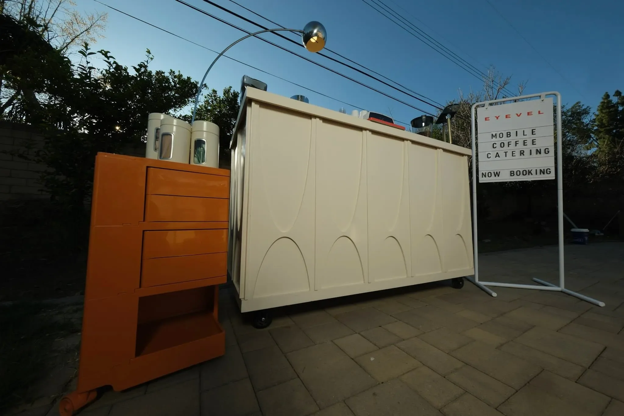A mobile coffee catering stand with equipment and signage that reads 'EyeLevel Mobile Coffee Catering Now Booking' set up outdoors on a tiled area with trees and power lines in the background.
