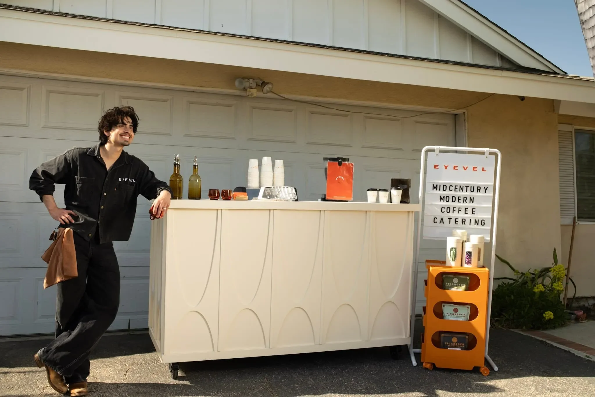 A man standing next to a mobile coffee stand outdoors, smiling, with various coffee supplies on the counter, and a sign advertising mid-century modern coffee catering.