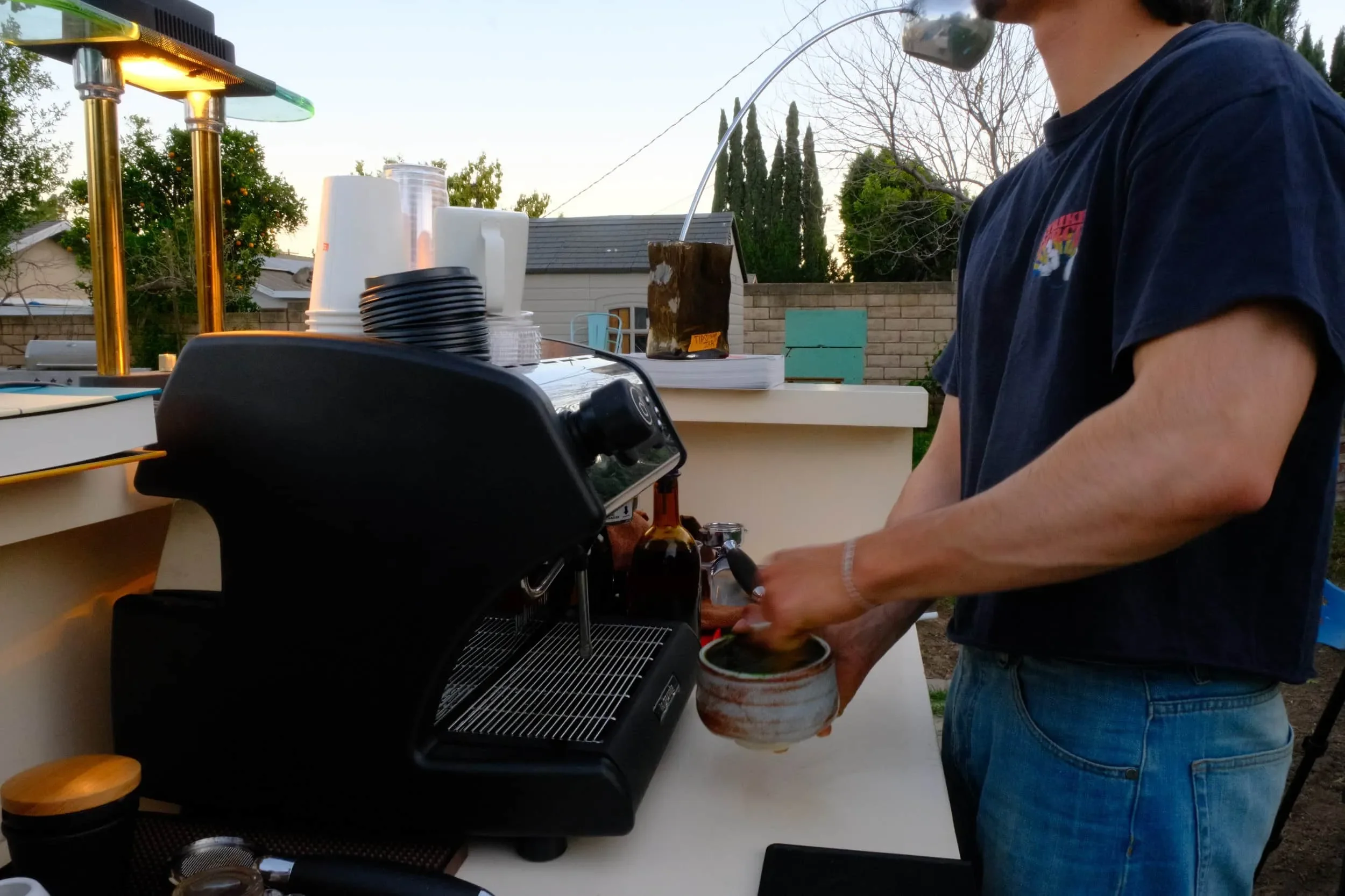 Person in a dark t-shirt and jeans making coffee or tea at an outdoor bar, with cups, plastic cups, napkins, and a catering chafing dish on the counter, trees, and a brick fence in the background.