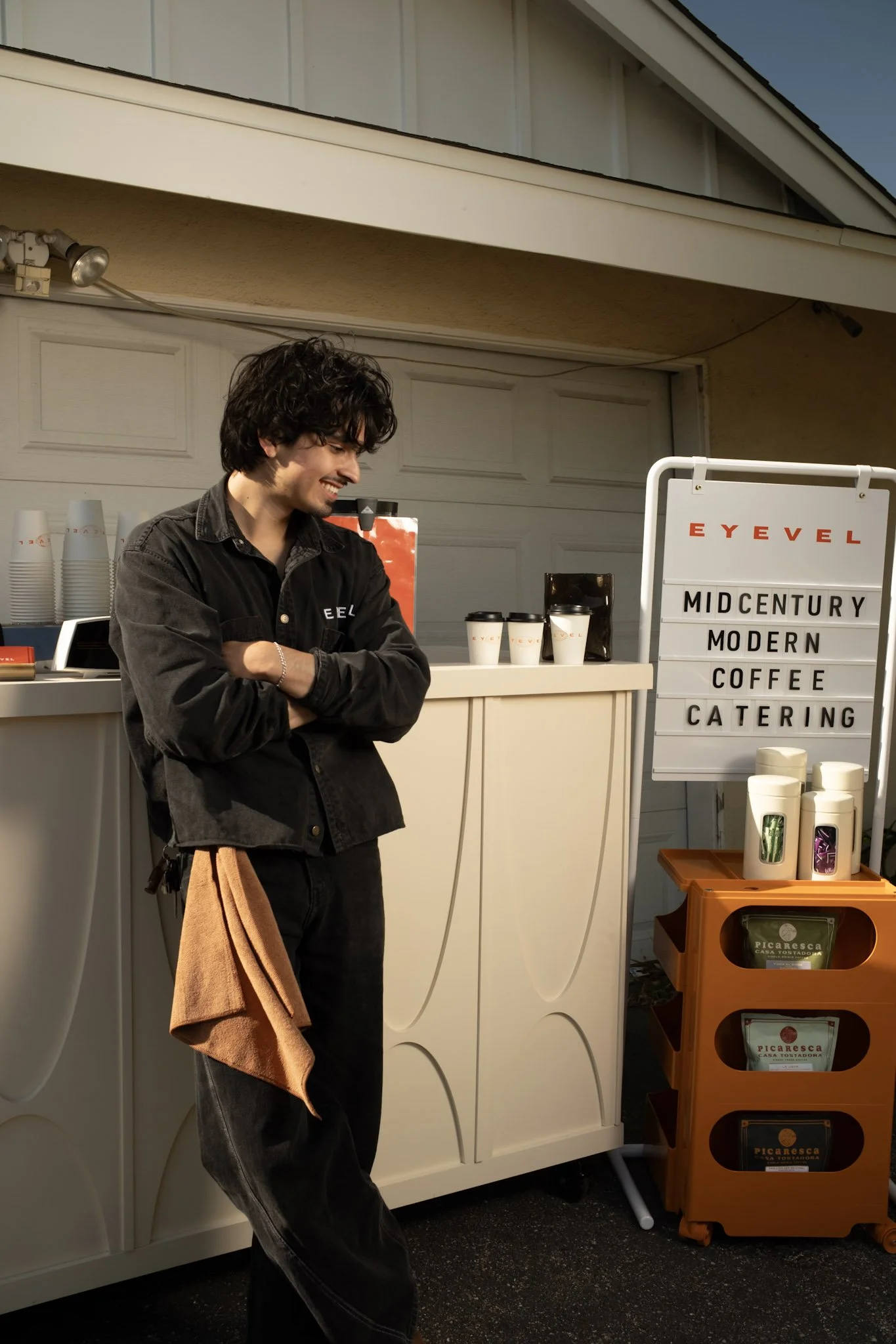 A young man with dark, curly hair and a black shirt stands with arms crossed, smiling outside a modern coffee stand with a sign that reads 'Mid Century Modern Coffee Catering.'