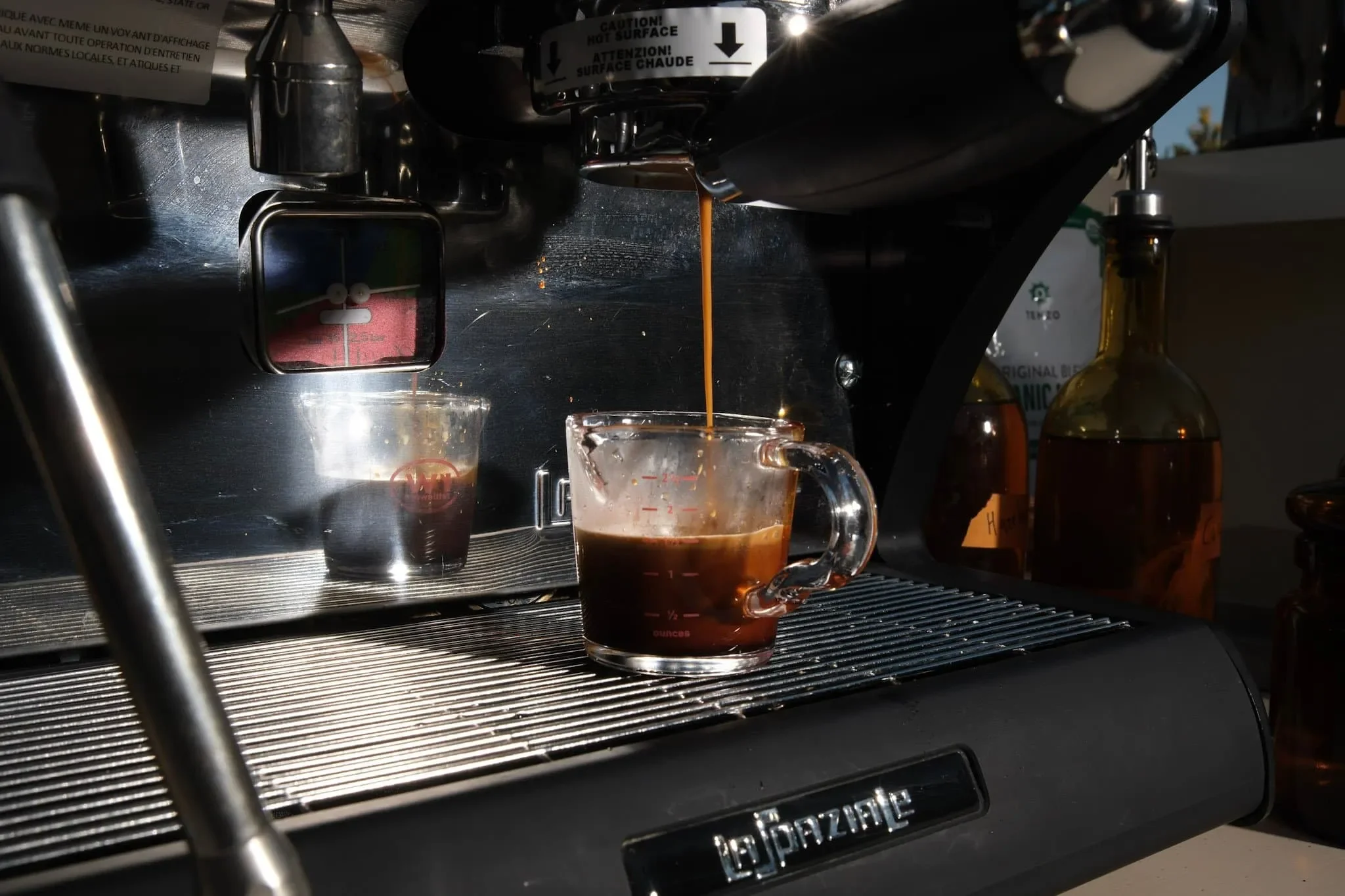 Close-up of a coffee machine dispensing hot espresso into a glass cup, with other bottles in the background.