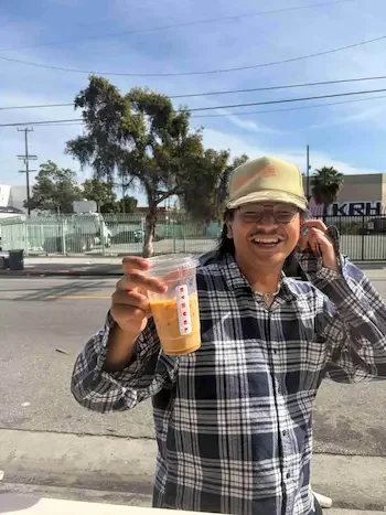 Person smiling and holding a plastic cup with a beverage, wearing a baseball cap and plaid shirt, standing outdoors on a sunny day.