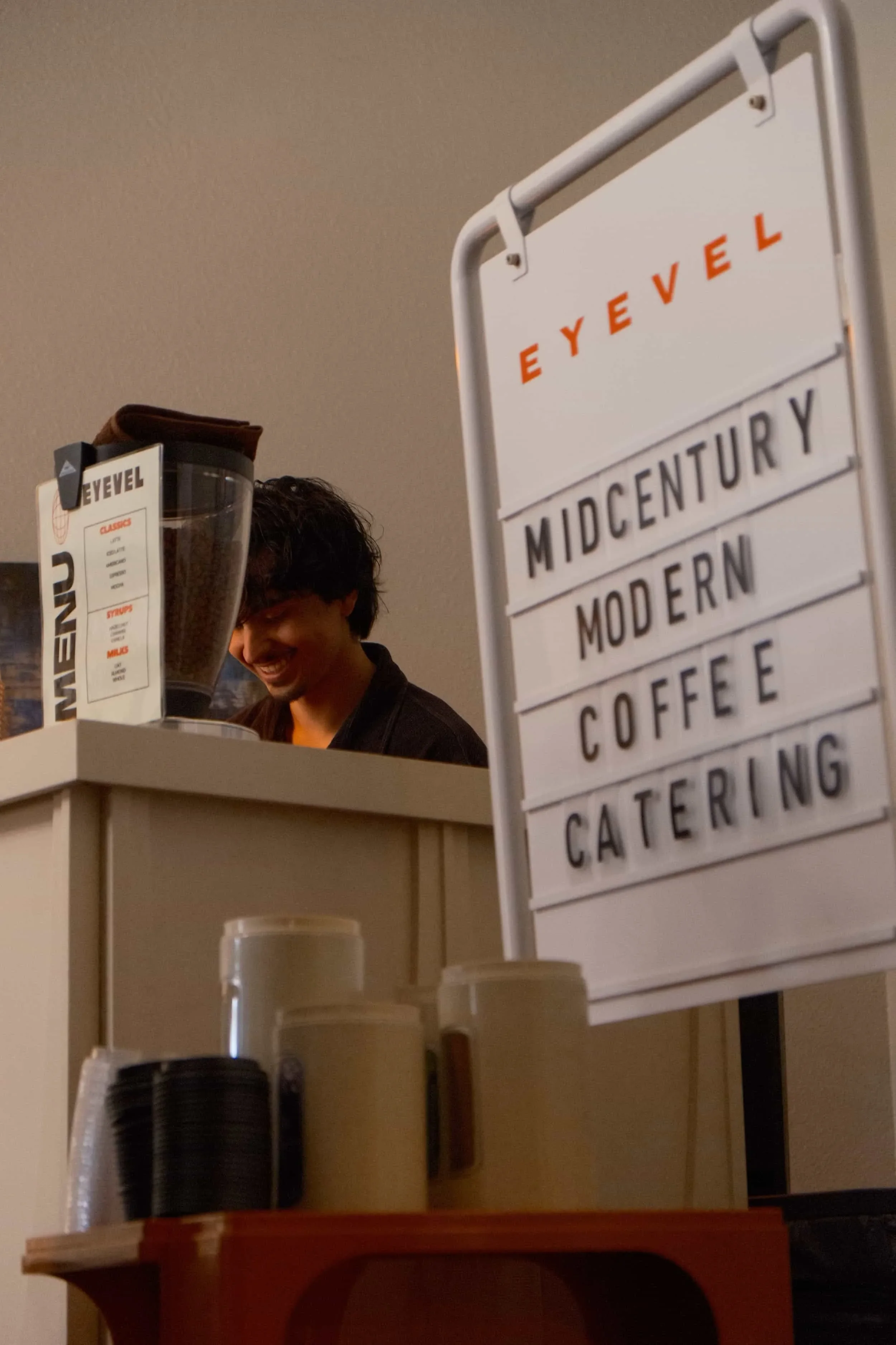 A person smiling behind a coffee counter with a white board indicating coffee, catering, and baking services. Coffee cups are visible in the foreground.