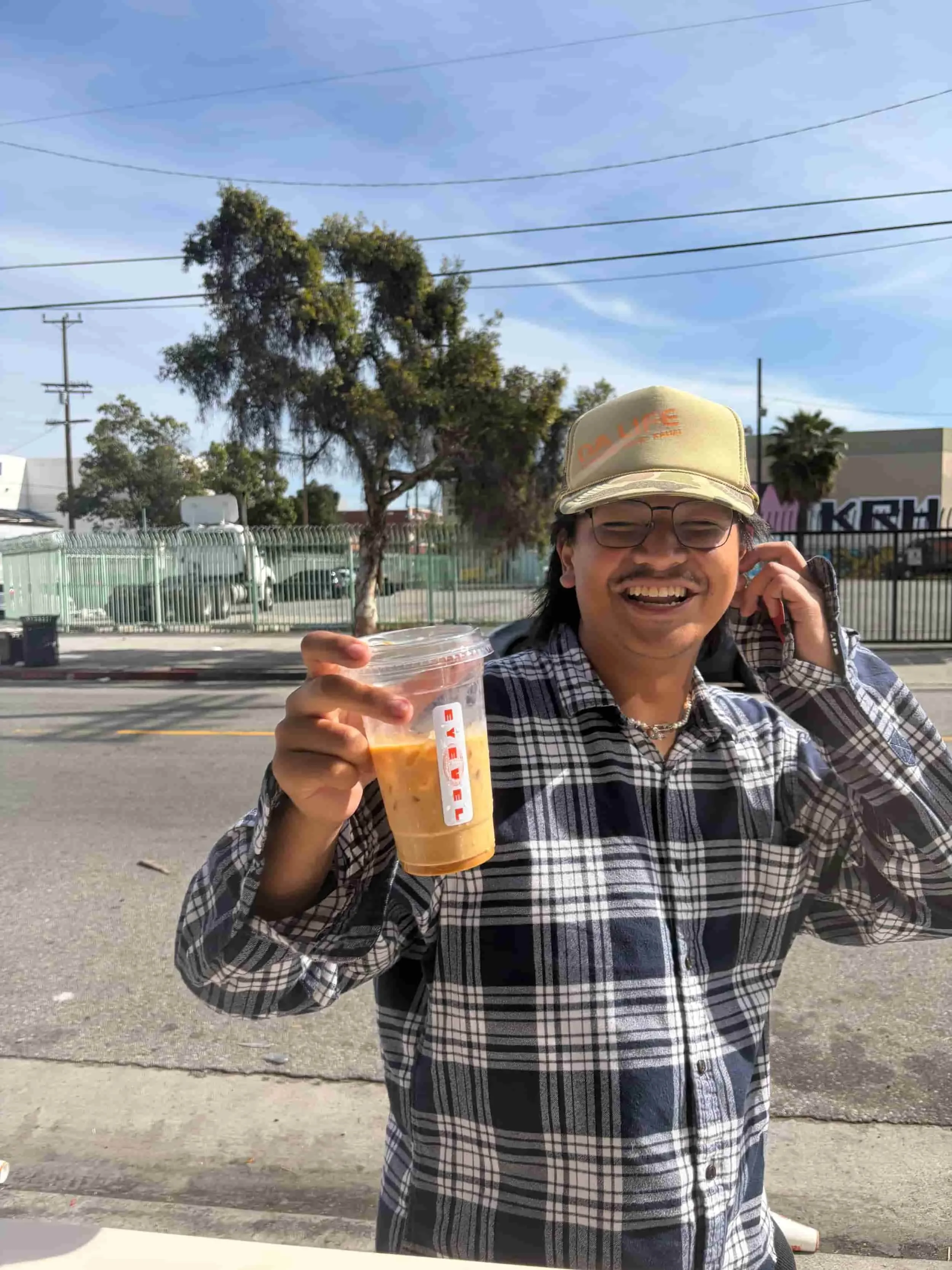 A man with glasses and a cap smiling and holding a large iced coffee cup outdoors on a sunny day, with trees, power lines, and buildings in the background.