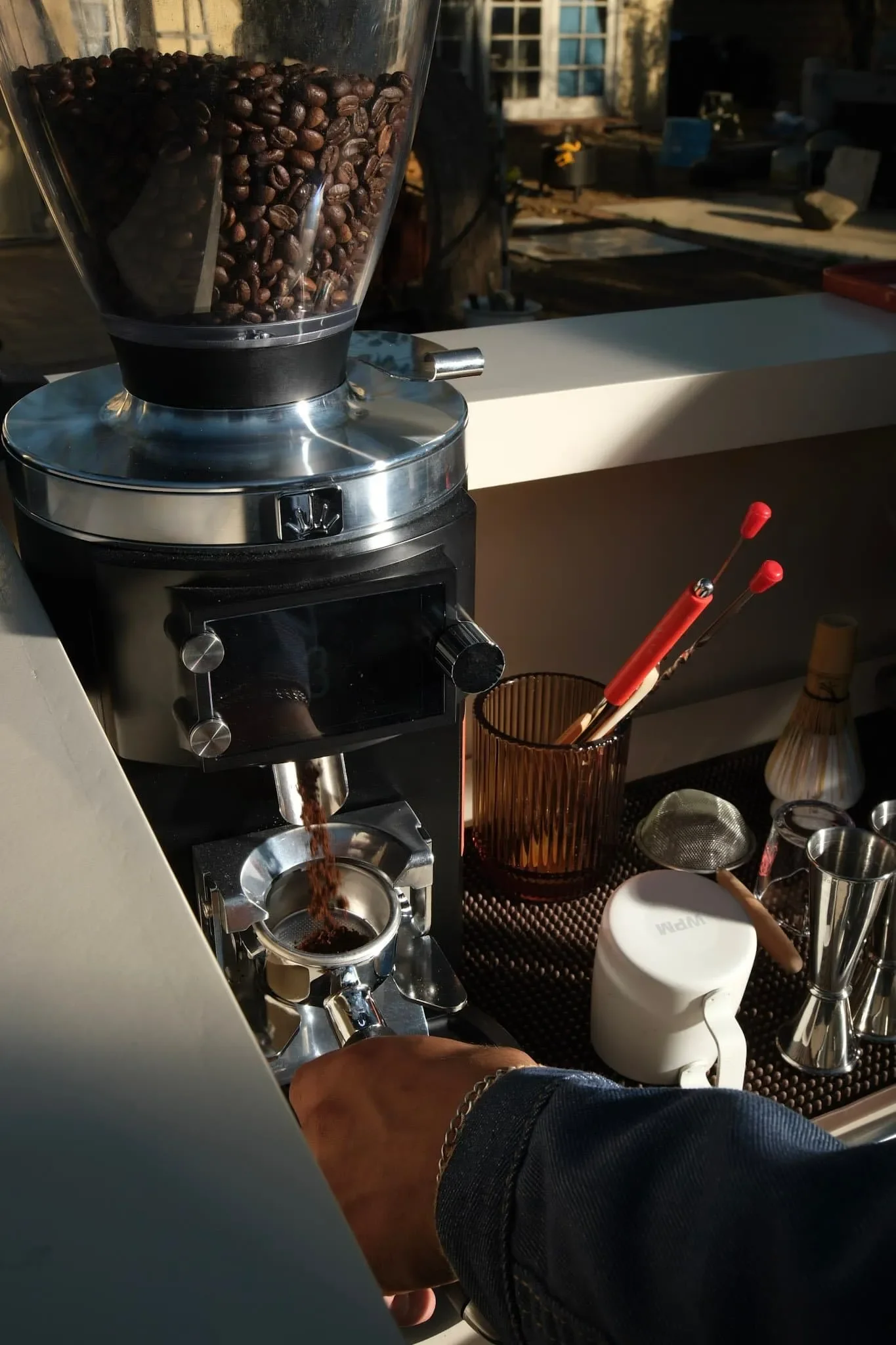Close-up of a coffee grinder with dark roasted coffee beans inside, grinding coffee into a cup below. The setting appears to be an outdoor café or coffee station with various utensils and equipment on the table.