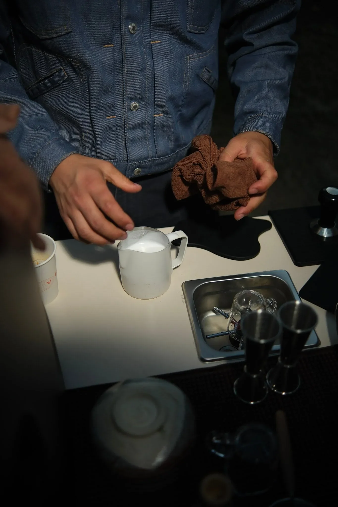 Person wearing a denim jacket holding a brown cloth, standing at a counter with a white pitcher, coffee mugs, and barista equipment.