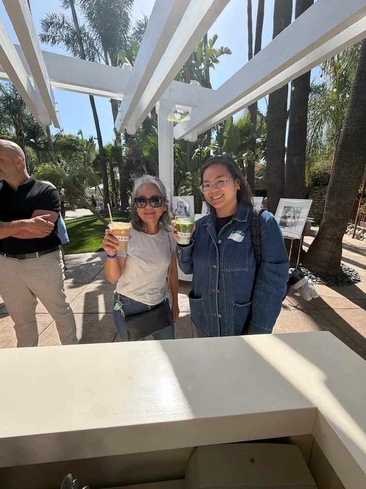 Two women smiling and holding colorful cups of ice cream or frozen treats outdoors on a sunny day, with palm trees and greenery in the background.