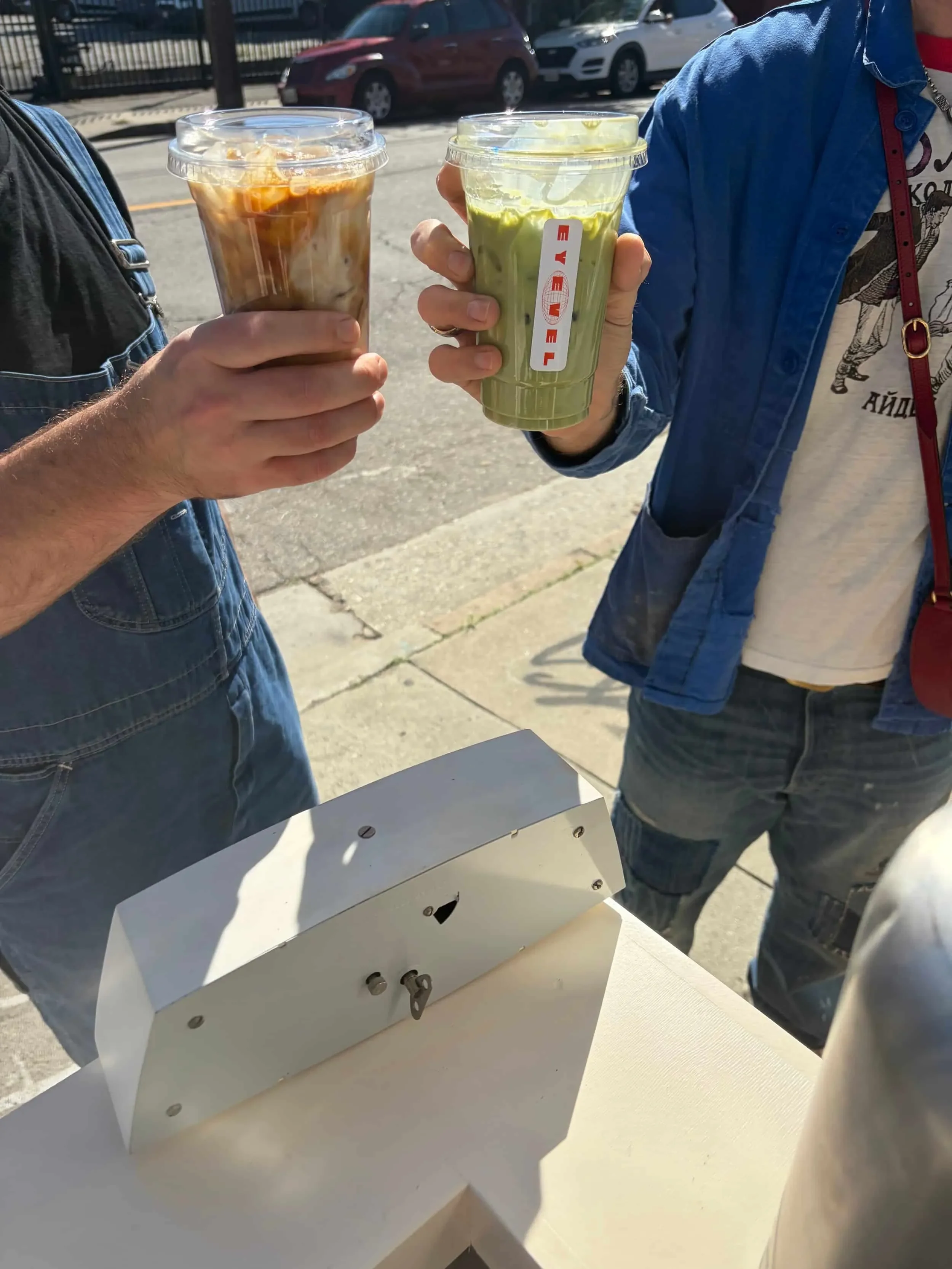 Two people holding iced coffee drinks, one with a brown caramel-colored iced coffee and the other with a green iced matcha latte, outdoors in sunlight.