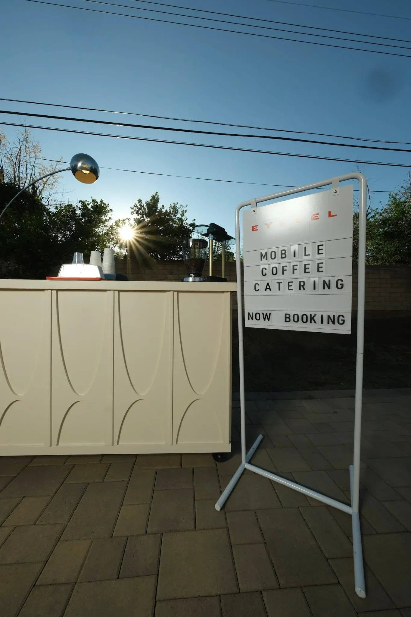 Outdoor scene with a white stand that has black letters and words advertising a mobile coffee catering service now booking. Behind the stand is a beige decorative wall, and on top of the wall are coffee supplies including a coffee grinder, cups, and 