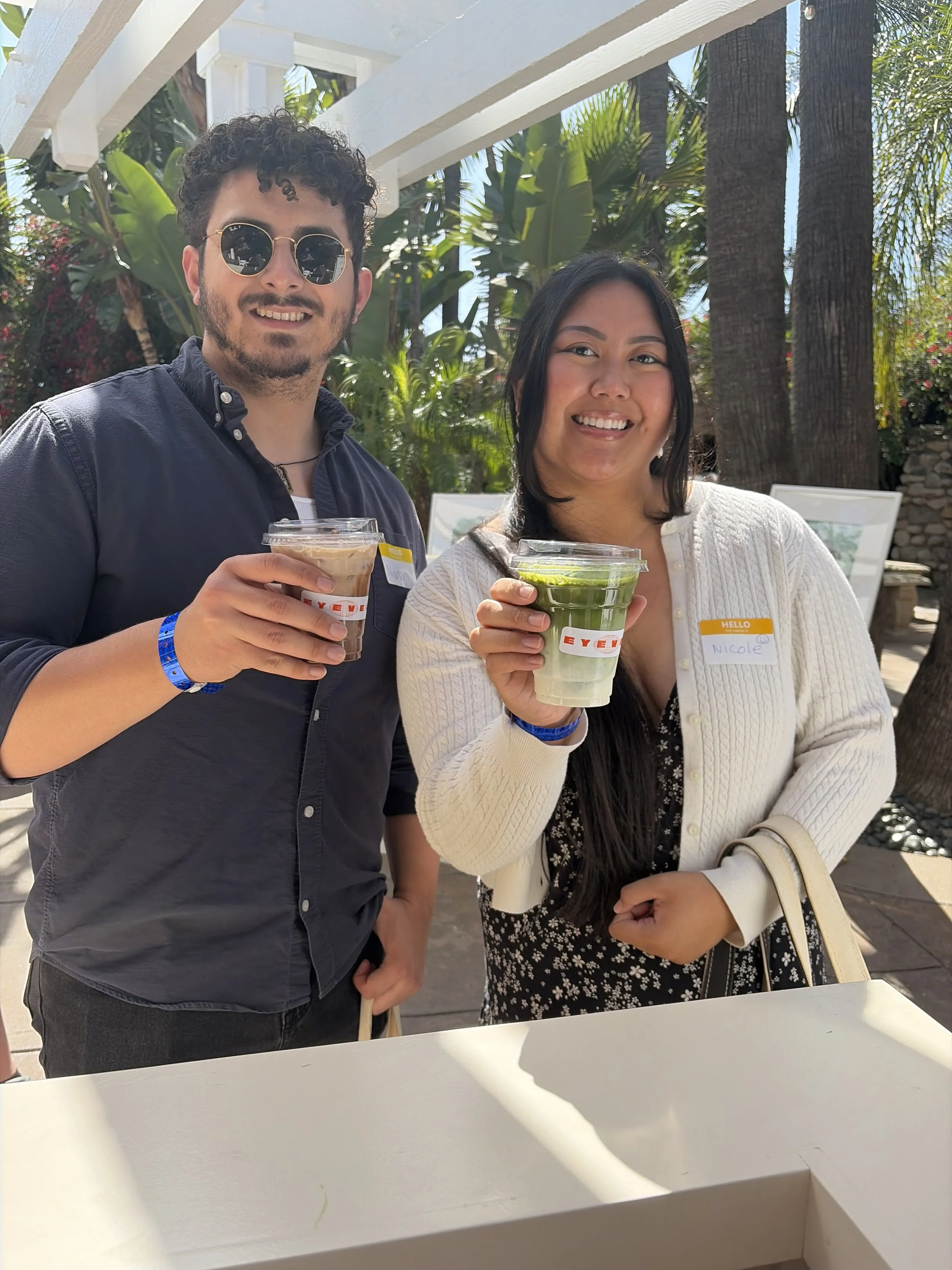 Two young adults, a man with curly hair, sunglasses, and a blue wristband, and a woman with long dark hair, a cream-colored cardigan, and a name tag reading "Nicole," are smiling and holding plastic cups with drinks, standing outdoors with palm trees